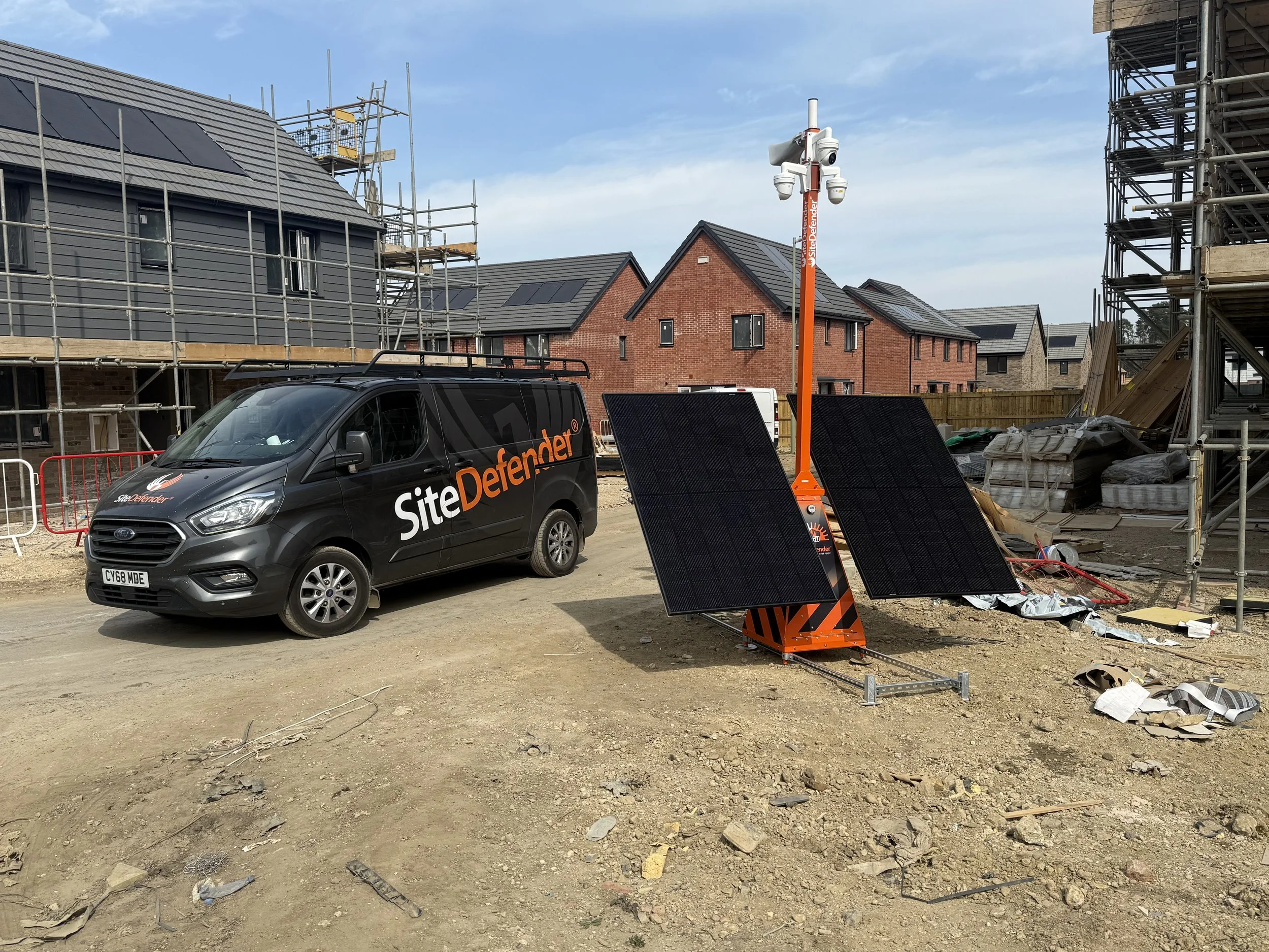 Construction site with a grey van labeled 'Site Defender', solar-powered surveillance camera, and solar panels on the ground, surrounded by partially built houses and scaffolding.