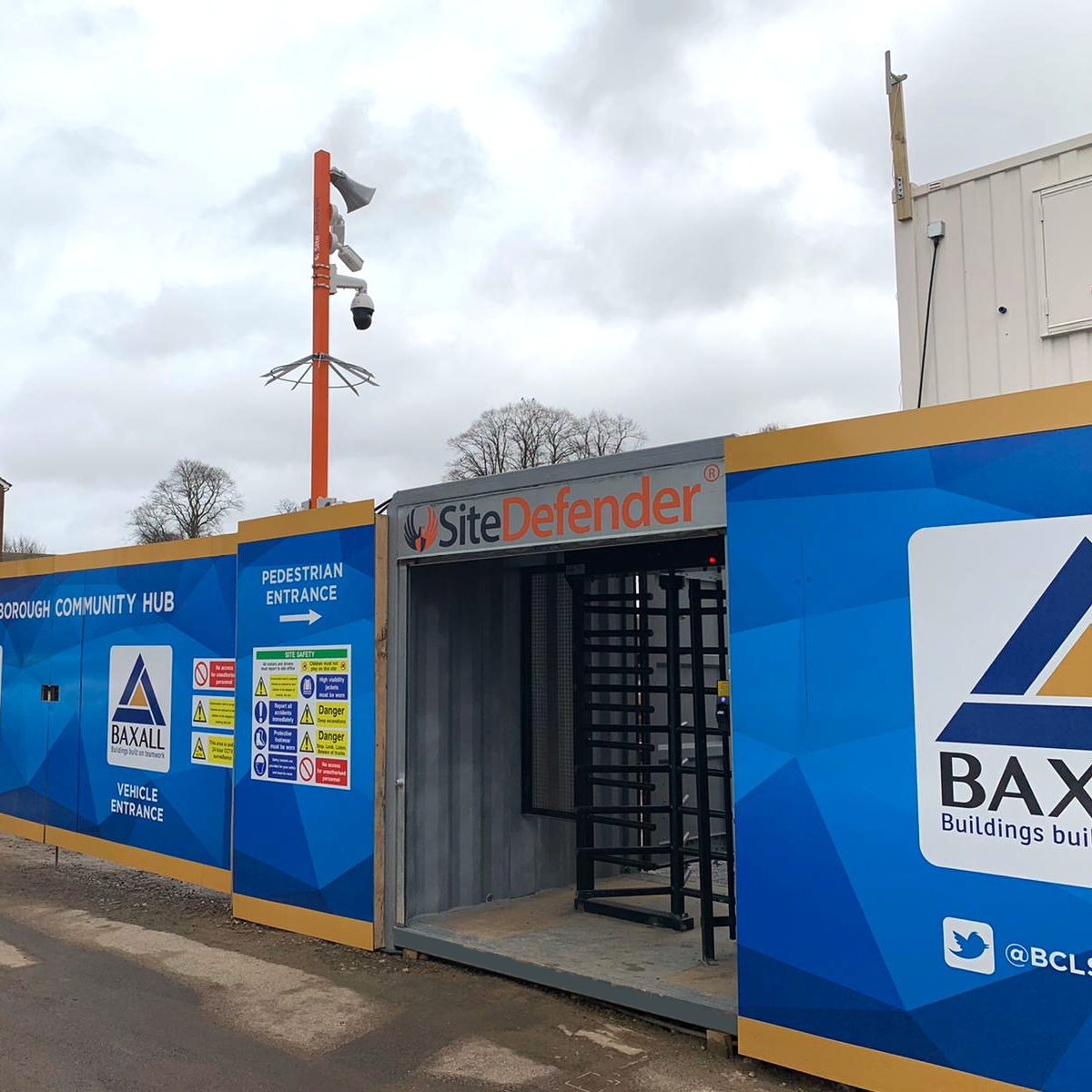 Construction site with blue and yellow fencing, a pedestrian entrance with signs, and a Site Defender security portal. There is a security camera mounted on a red pole, and the sky is cloudy with leafless trees in the background.