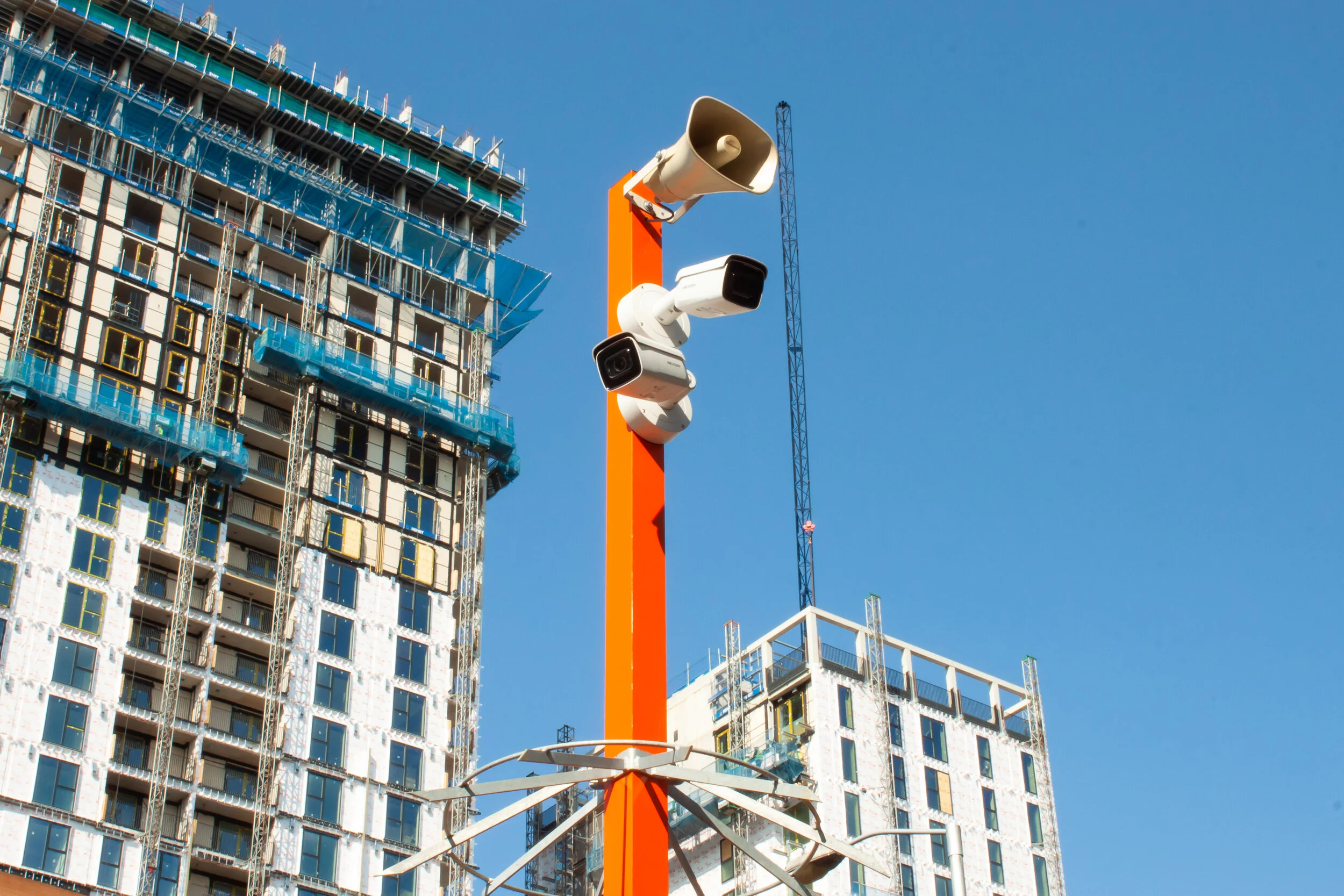 Construction site with a tall orange pole holding multiple security cameras and a loudspeaker, with a building under construction in the background and a clear blue sky.