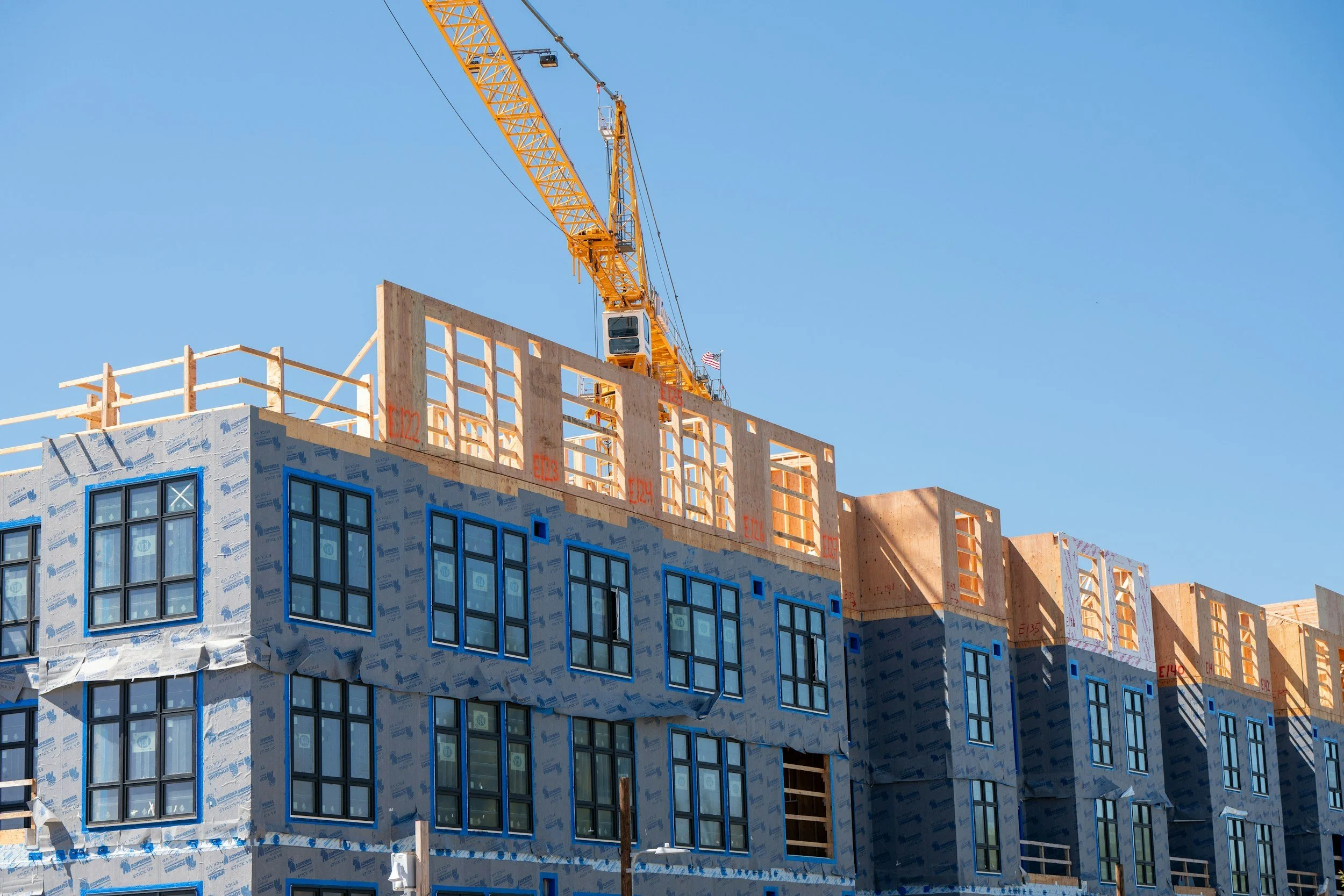 Construction site of a multi-story building with windows installed, building frame partially unfinished, and a large yellow crane lifting materials against a clear blue sky.
