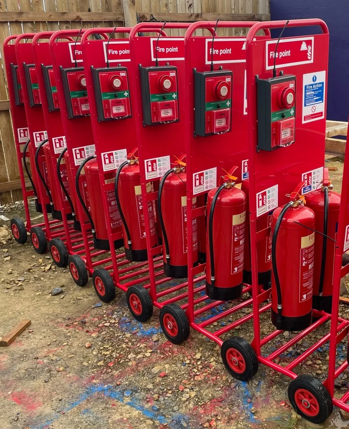 A fire safety station with two red fire extinguishers, a fire alarm, and a fire plan on a red stand outside a construction site, surrounded by a metal fence.