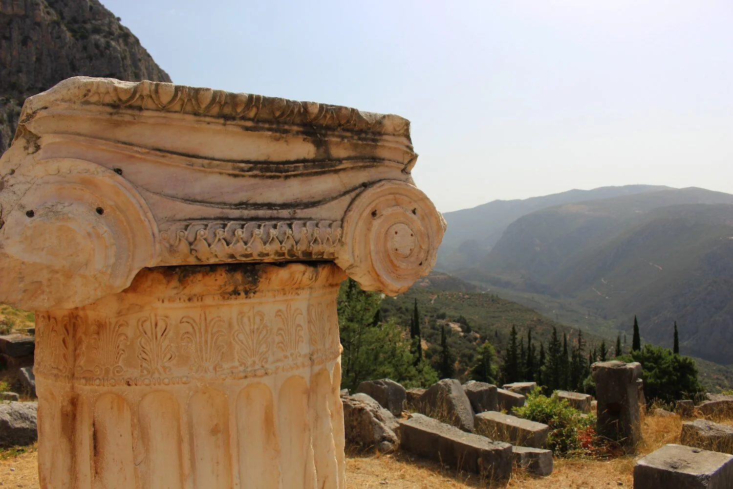 A close-up photograph of a Delphic temple column with carved designs of acanthus leaves and geometric shapes, with a view of the valley beneath Delphi in the background.