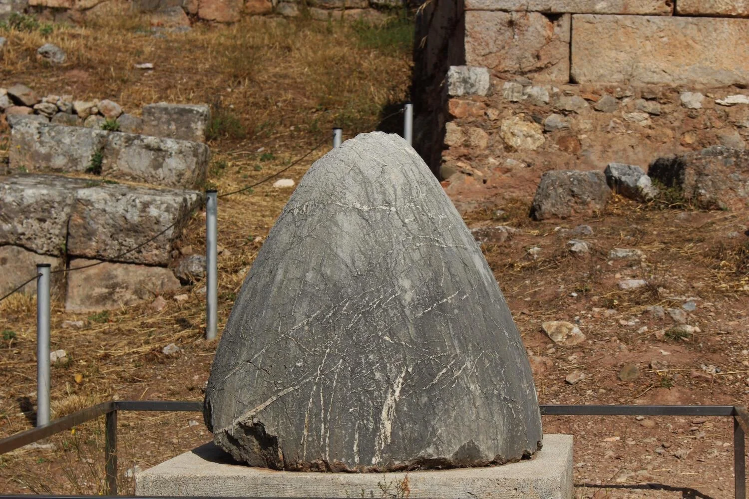 A photograph of a grey navel stone, the Omphalos, on a concrete slab surrounded by a metal railing.