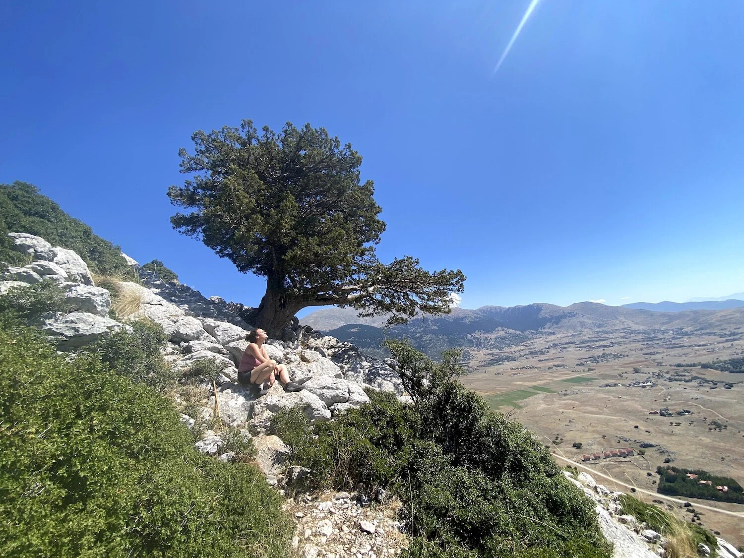 A photograph of me, Ekstasy, sitting on the rocks outside of the Corycian Cave, looking up at the sky with a view of the Livadi valley in the background. Photograph taken by Erin Bernstein.