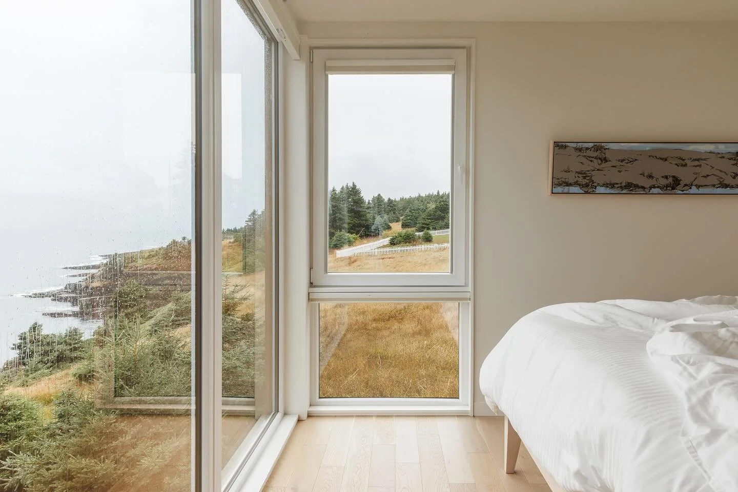 Rainy mornings in Amherst Cove. Corner windows in the primary bedroom frame a sweeping view of the coastline.

Inside, local artistry and craftsmanship are on display with artwork by @barbarahouston_artstudio and a custom bed designed and built by @b
