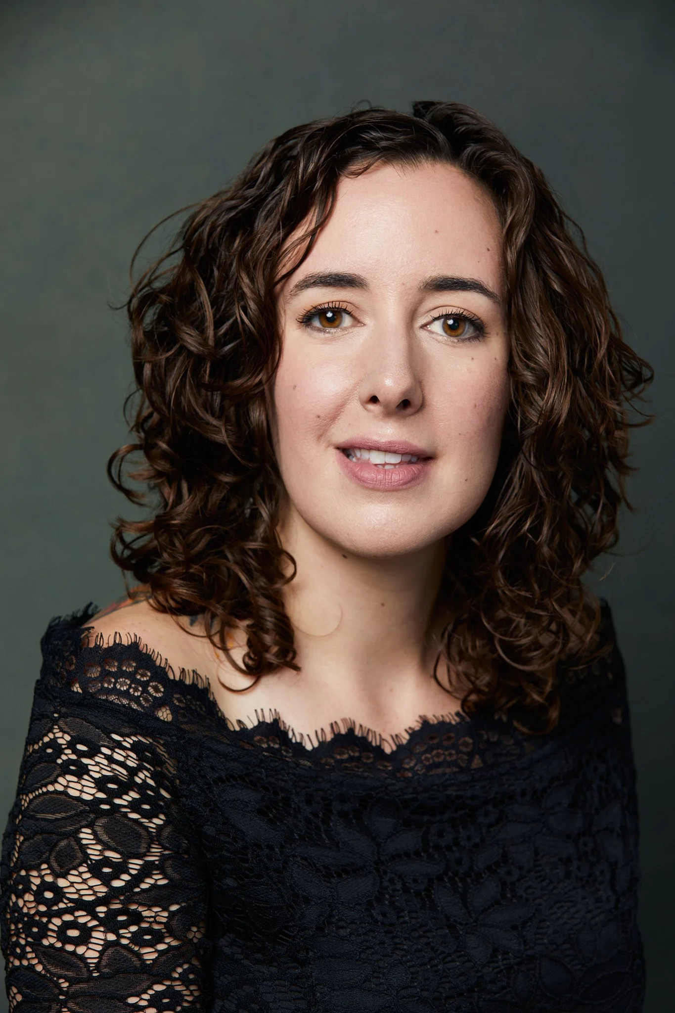 Portrait of a woman with curly brown hair, fair skin, wearing a black lace top, against a dark green background.