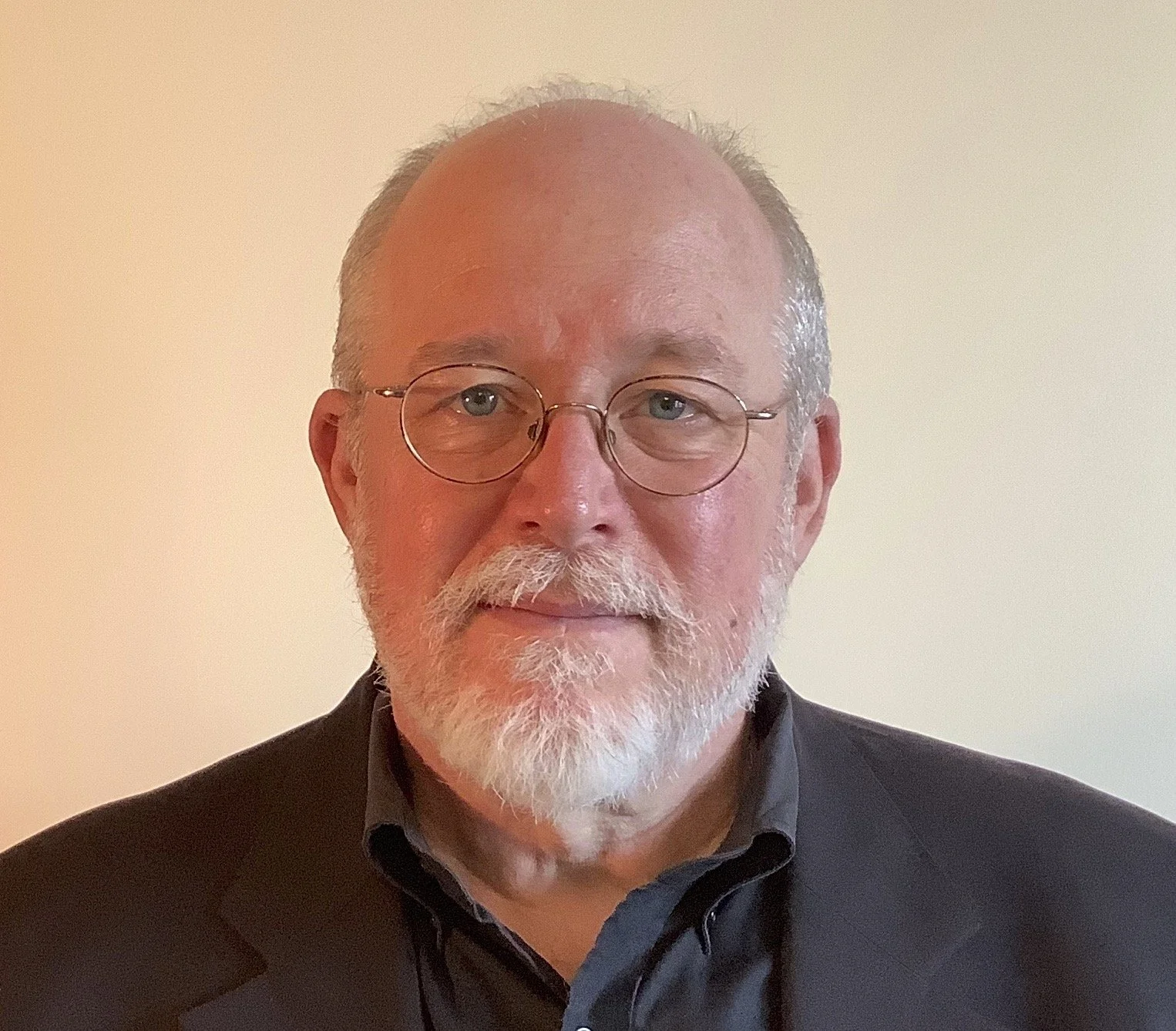 A middle-aged man with light skin, glasses, a white beard, and a receding hairline wearing a black blazer over a dark shirt, standing against a plain beige wall.
