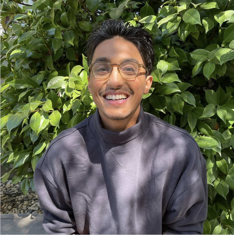 A young man smiling outdoors, wearing glasses and a gray sweatshirt, standing in front of lush green leaves.