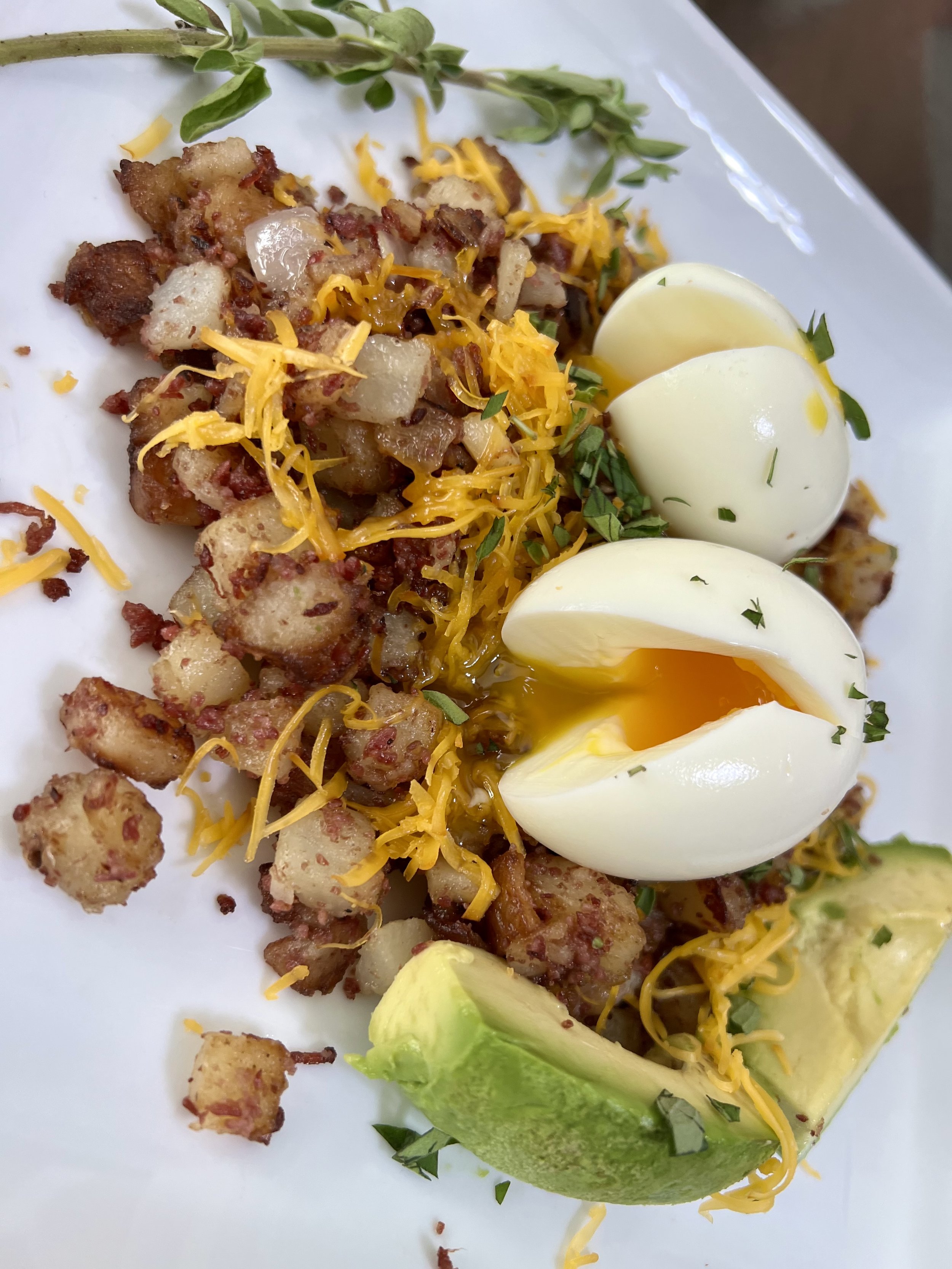 A hearty breakfast plate featuring crisp potato and corned beef hash, topped with shredded cheddar cheese, fresh avocado slices, and two halved soft-boiled eggs with warm, runny yolks.