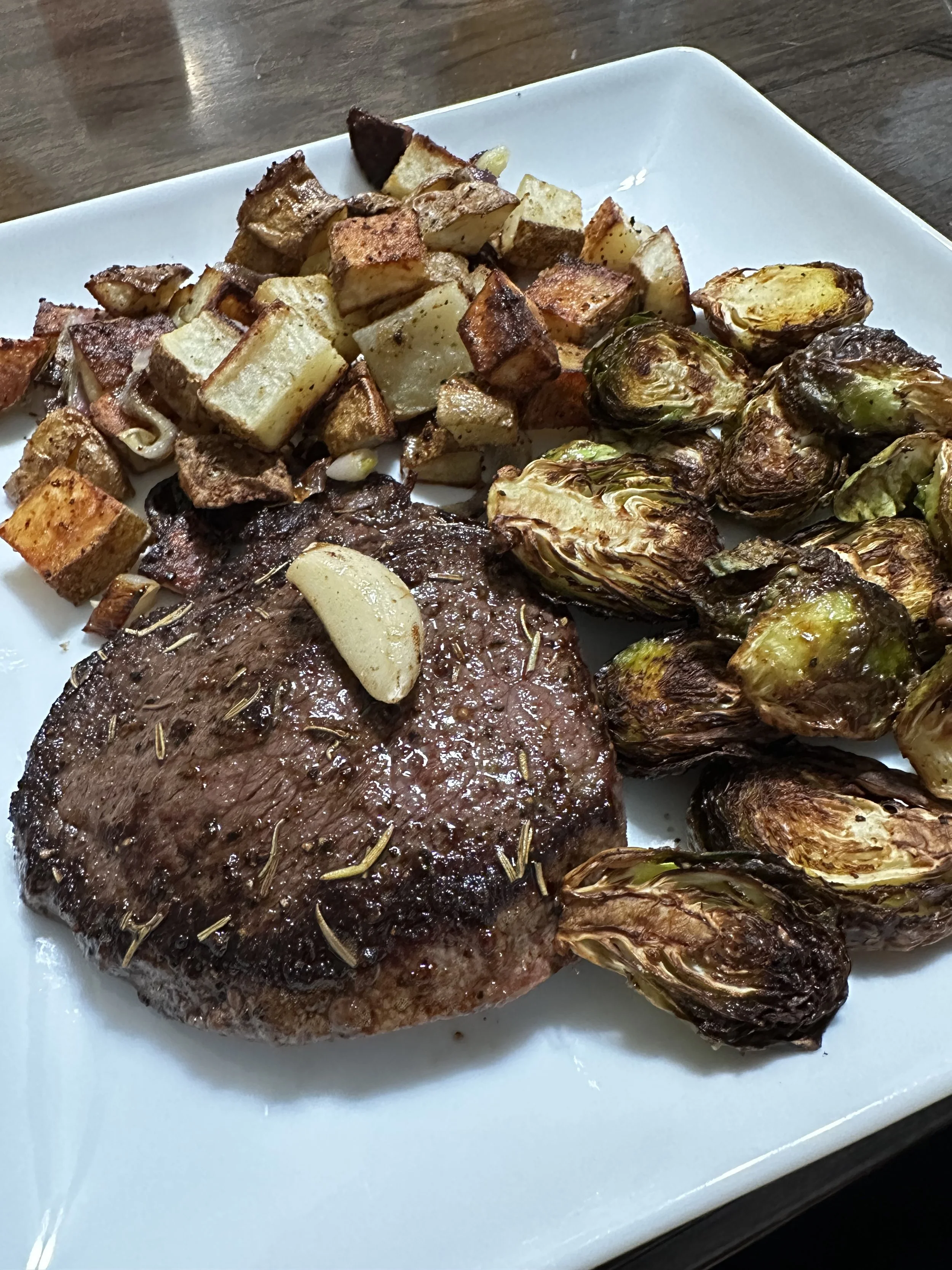 A juicy, seared sirloin steak topped with a clove of roasted garlic, served on a white plate with crisp, golden roasted potato cubes and caramelized roasted brussels sprouts.