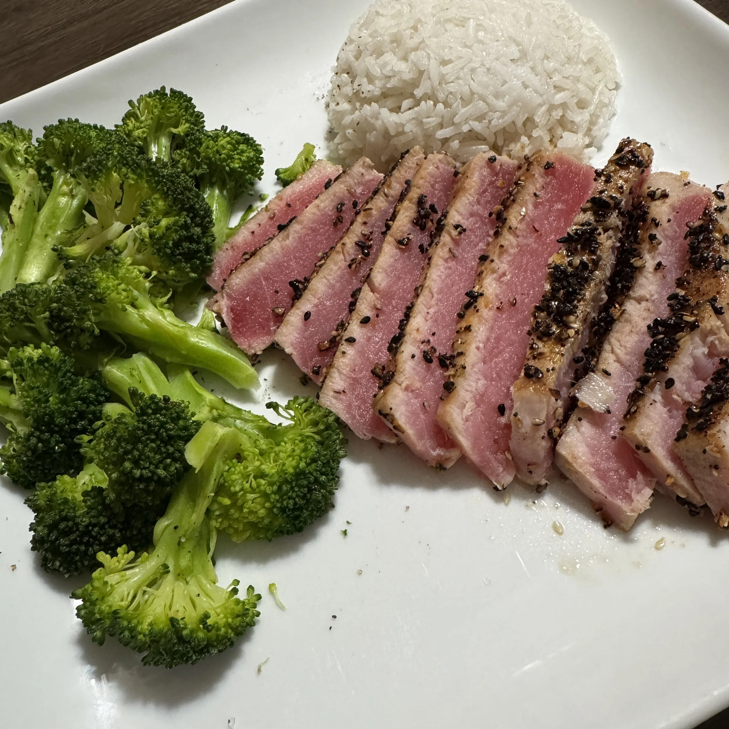 A white plate featuring sliced, rare seared ahi tuna with a toasted sesame seed crust, showcasing a vibrant pink center. It is served alongside bright green steamed broccoli florets and a neatly molded dome of white jasmine rice.