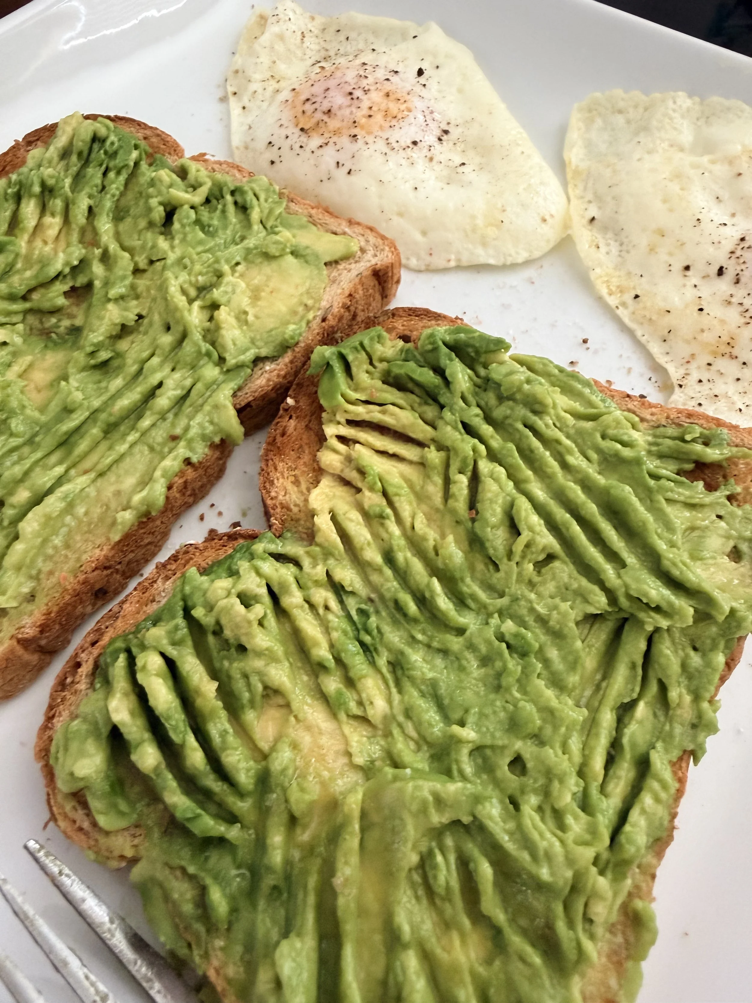 Sliced whole-grain toast layered with thick, bright green mashed avocado, topped with sliced hard-boiled eggs, red pepper flakes, and microgreens.