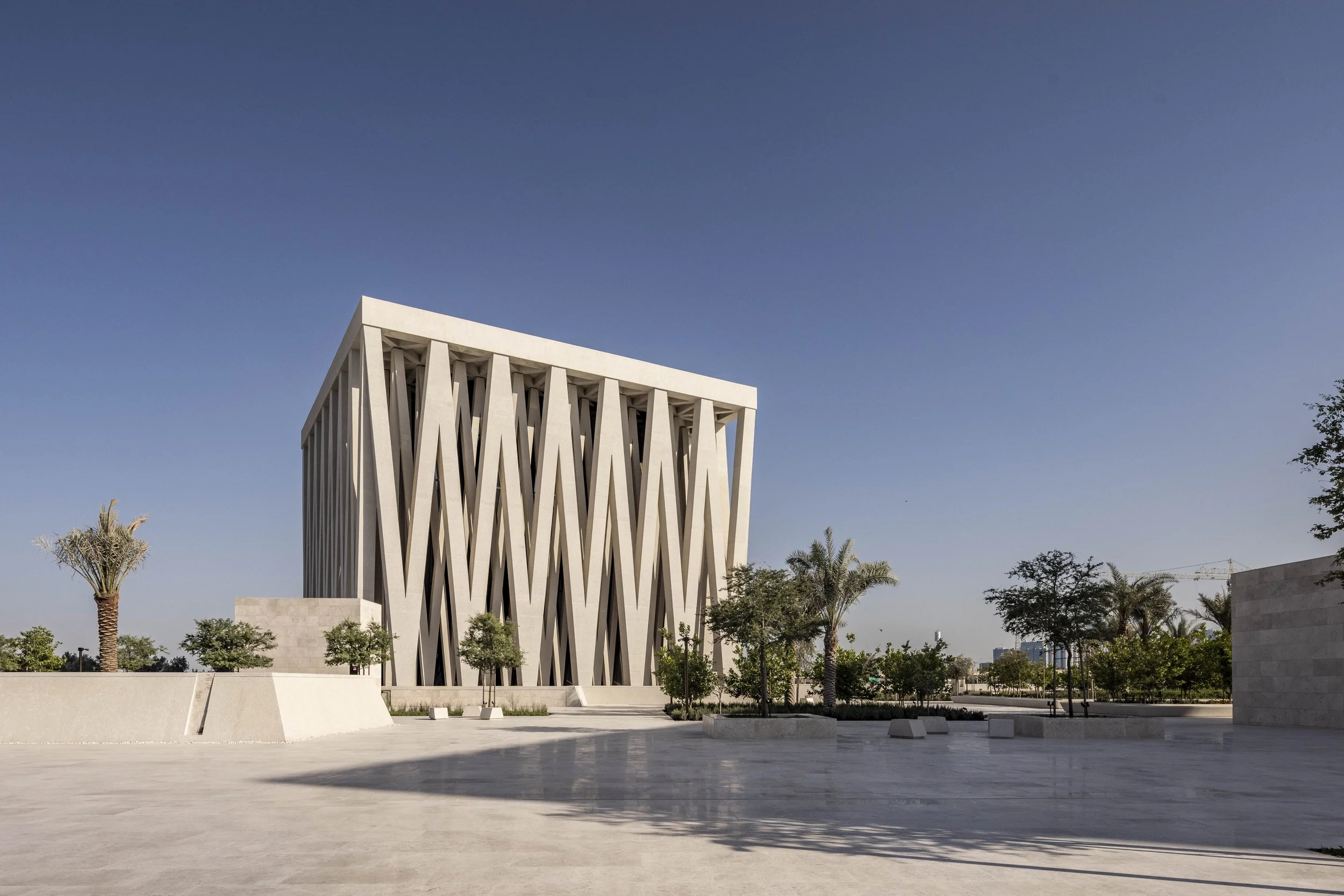 Moses Ben Maimon Synagogue with V-shaped columns on a stone plaza among palms, Abrahamic Family House, Abu Dhabi. Architect Adjaye Associates