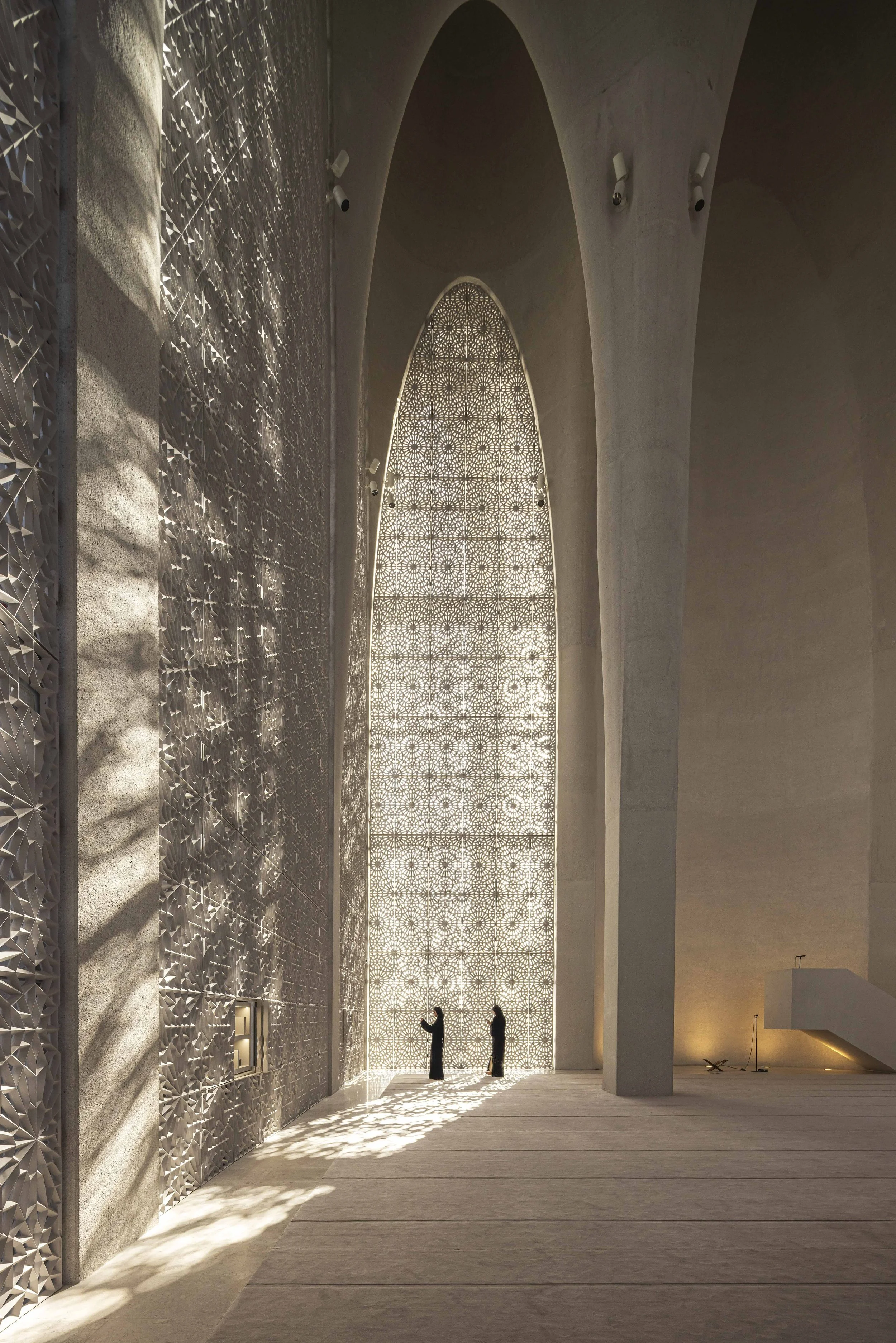 Two figures beneath a pointed arch and mashrabiya lattice window inside the Imam Ahmed Al-Tayeb Mosque, Abrahamic Family House, Abu Dhabi. Architect Adjaye Associates