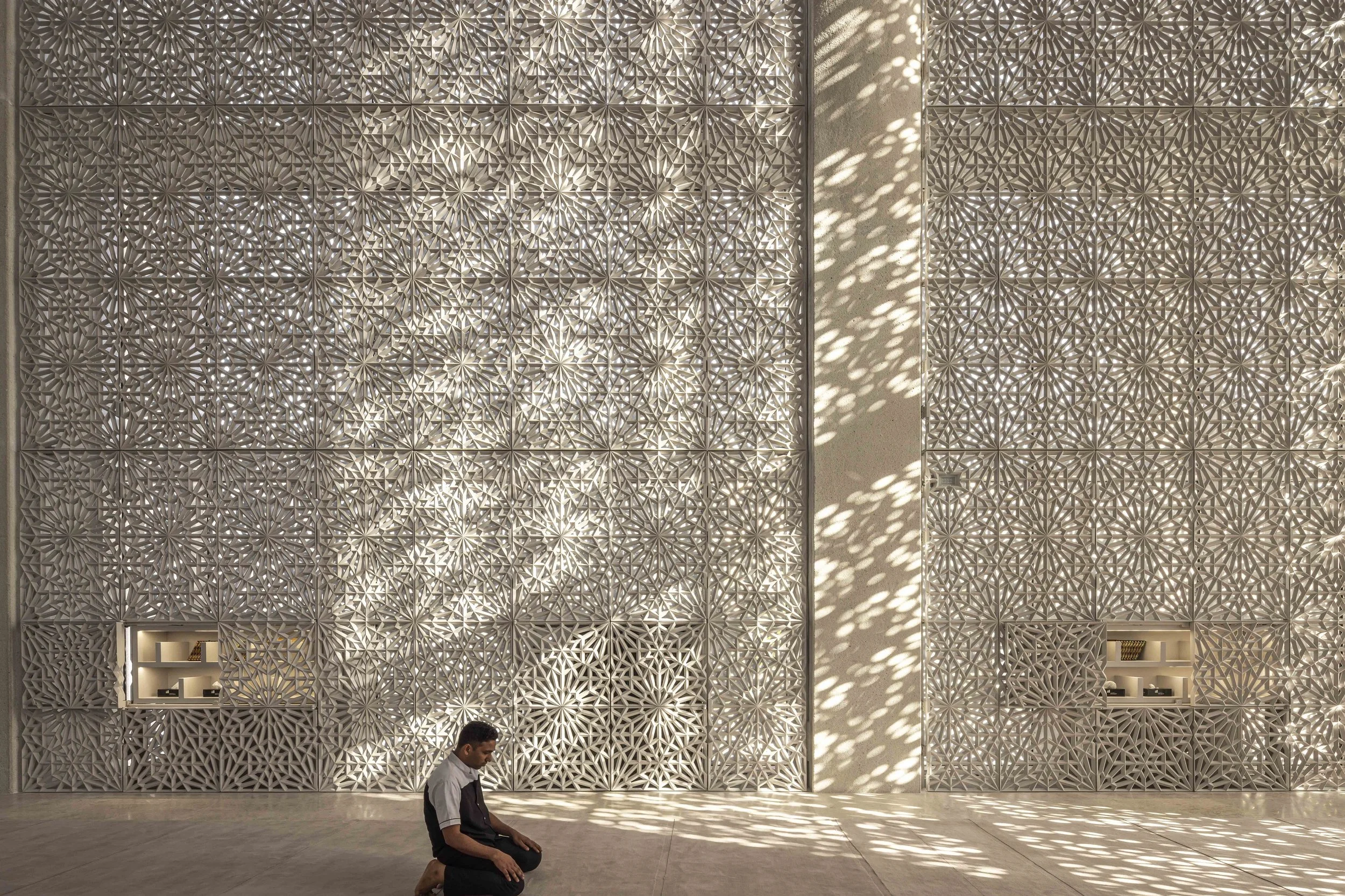 Man kneeling in prayer before the towering lattice wall of the Imam Ahmed Al-Tayeb Mosque, Abrahamic Family House, Abu Dhabi. Architect Adjaye Associates