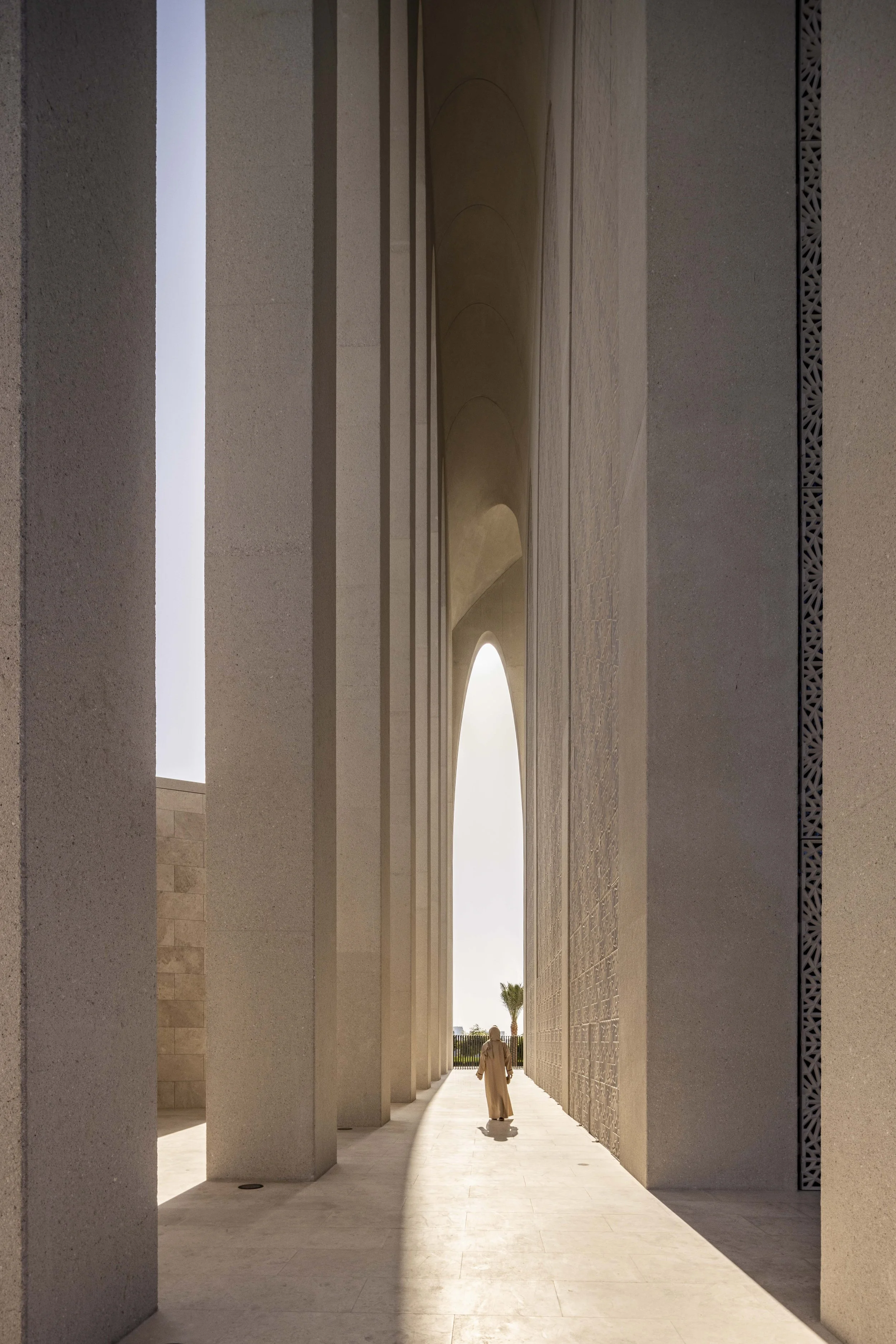 Figure in a cream thobe walking the colonnade of the Imam Ahmed Al-Tayeb Mosque toward a pointed arch, Abrahamic Family House, Abu Dhabi. Architect Adjaye Associates