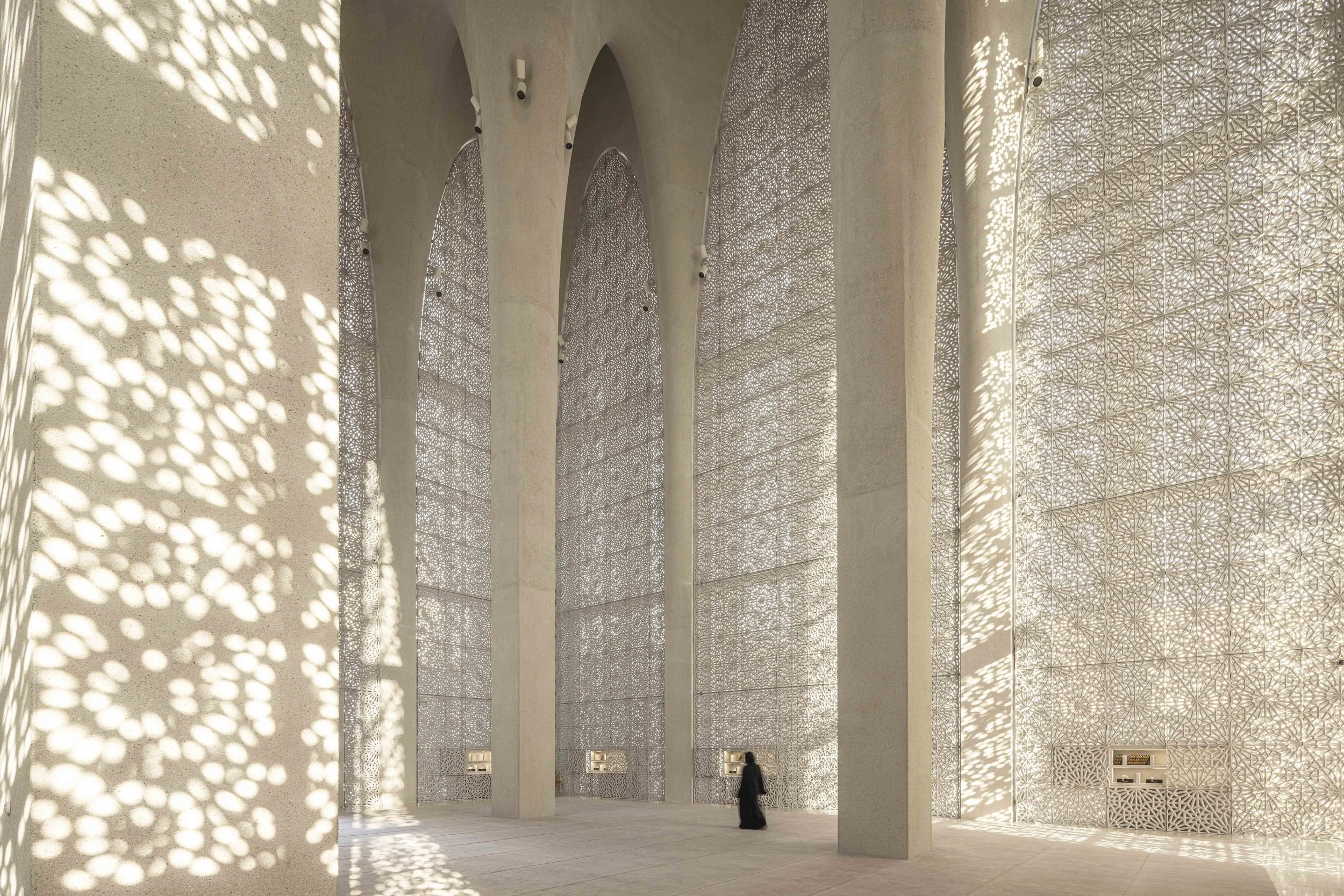 Light through lattice screens across tall arches and columns inside the Imam Ahmed Al-Tayeb Mosque, Abrahamic Family House, Abu Dhabi. Architect Adjaye Associates