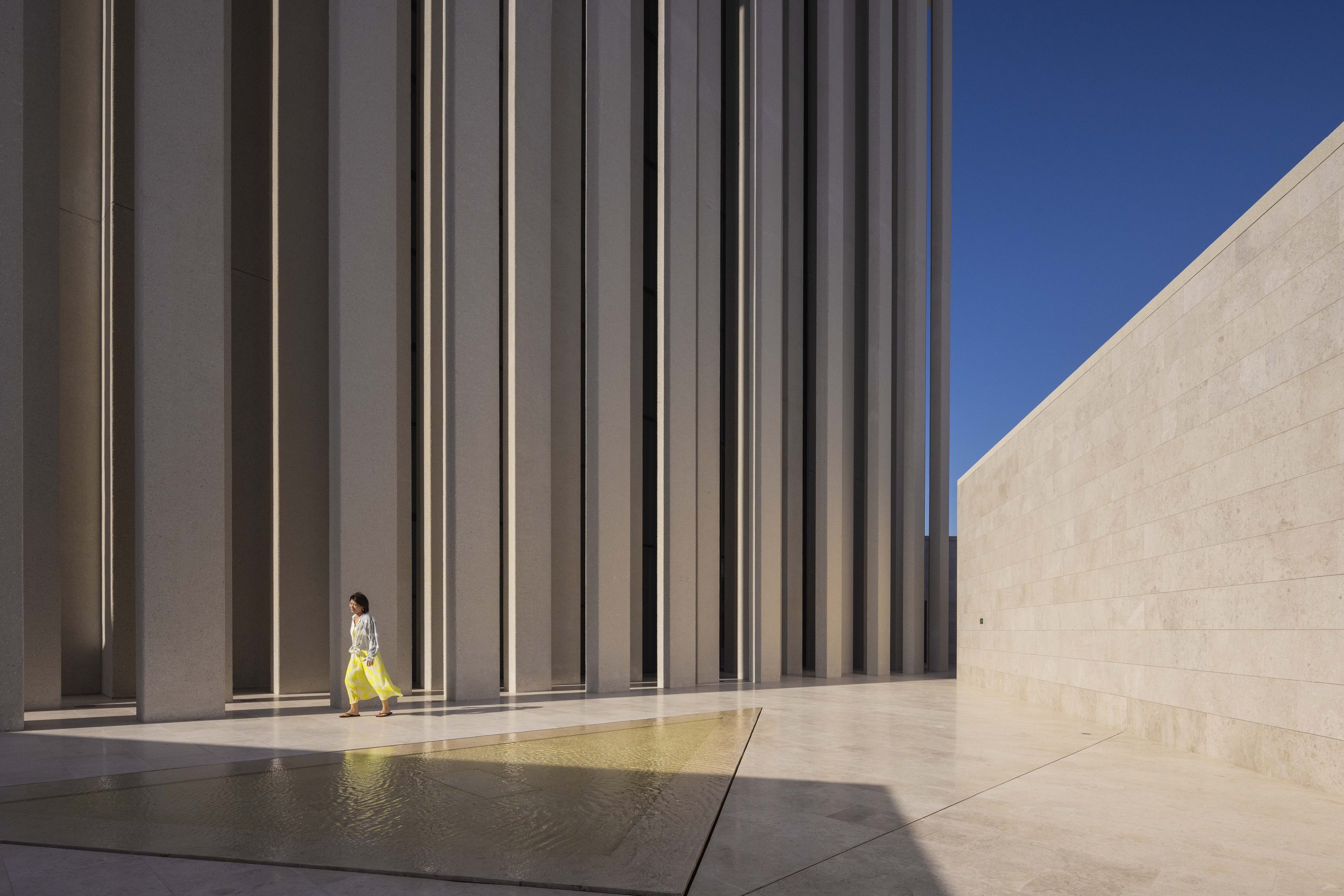 Woman in yellow crossing the courtyard beside the straight pillars of St. Francis Church, Abrahamic Family House, Abu Dhabi. Architect Adjaye Associates