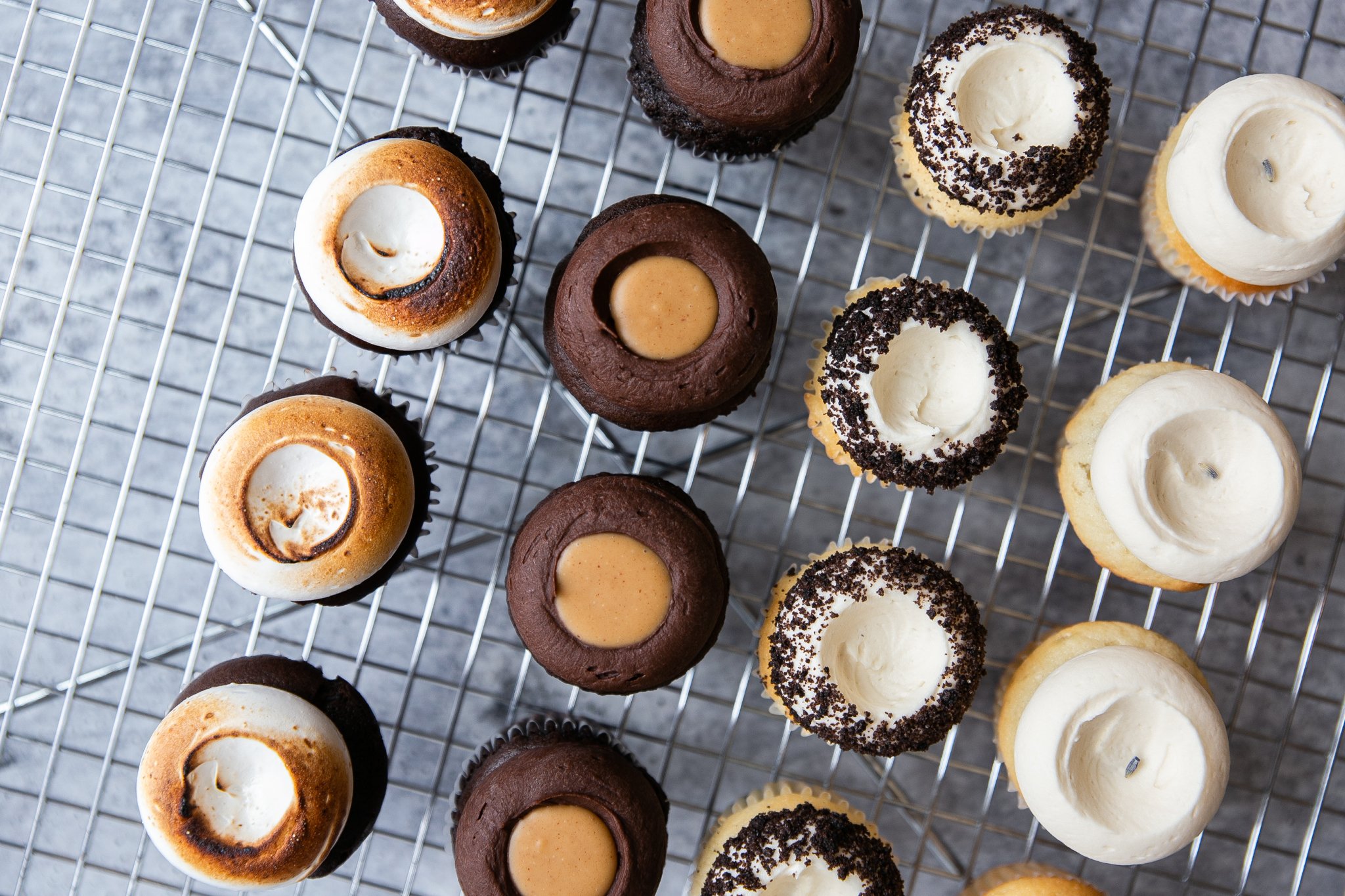  16 cupcakes are on a silver cooling tray. One row of s’mores cupcakes. One row of buckeye cupcakes. One row of cookies &amp; cream cupcakes. One row of white lavender cupcakes. 