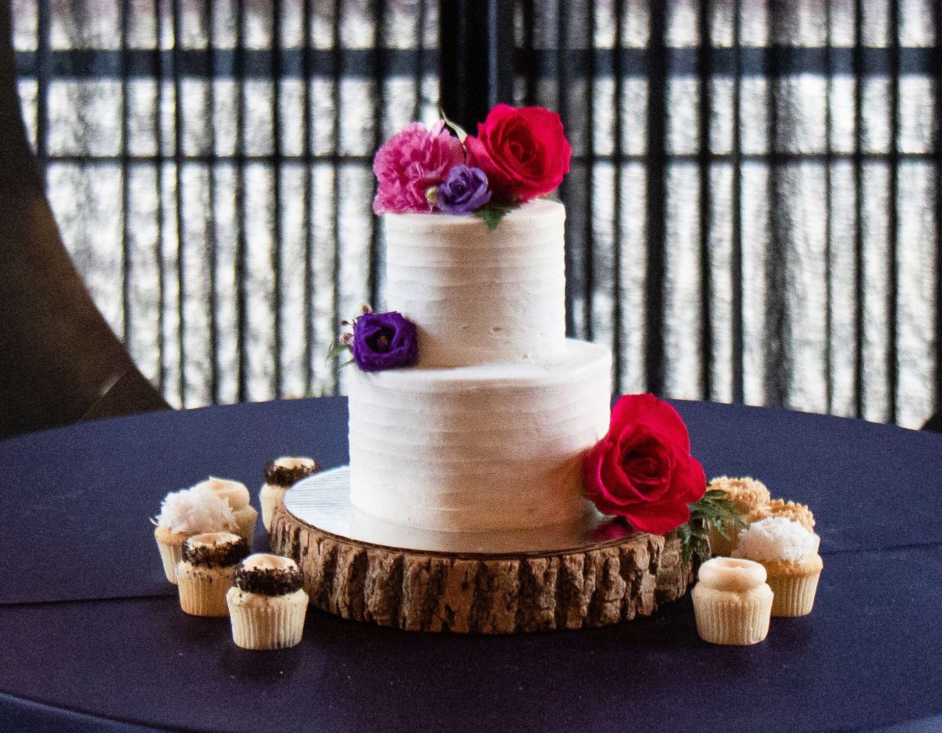  A white 2-tiered cake decorated with purple and pink flowers. A few cupcakes are displayed around the base of the cake. 