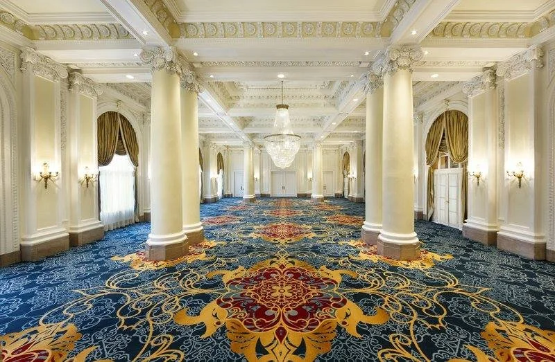 Spacious, elegant ballroom with white columns, ornate crown molding, chandeliers, and large windows with gold curtains. The room has a richly patterned blue, red, and gold carpet.