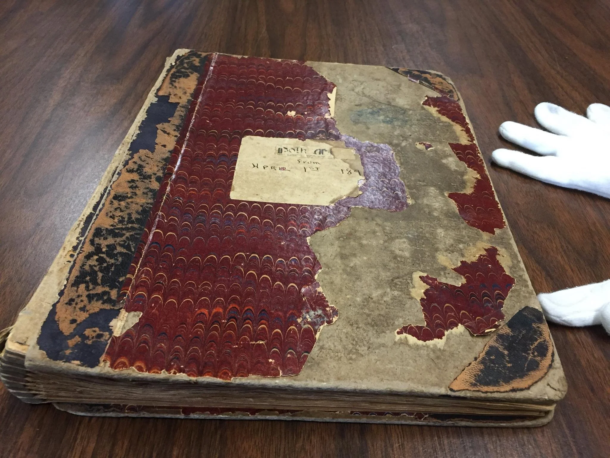 Old, damaged book with worn out, decorative marbled cover and a partially torn label on the front, lying on a wooden surface.