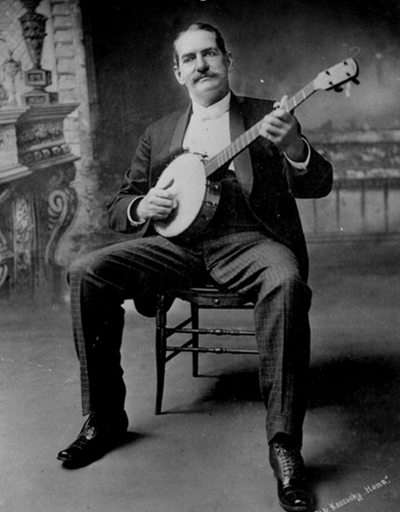 A black and white photo of a man sitting on a wooden chair, playing a banjo, wearing a suit and tie, with a background that appears to be a painted studio backdrop.