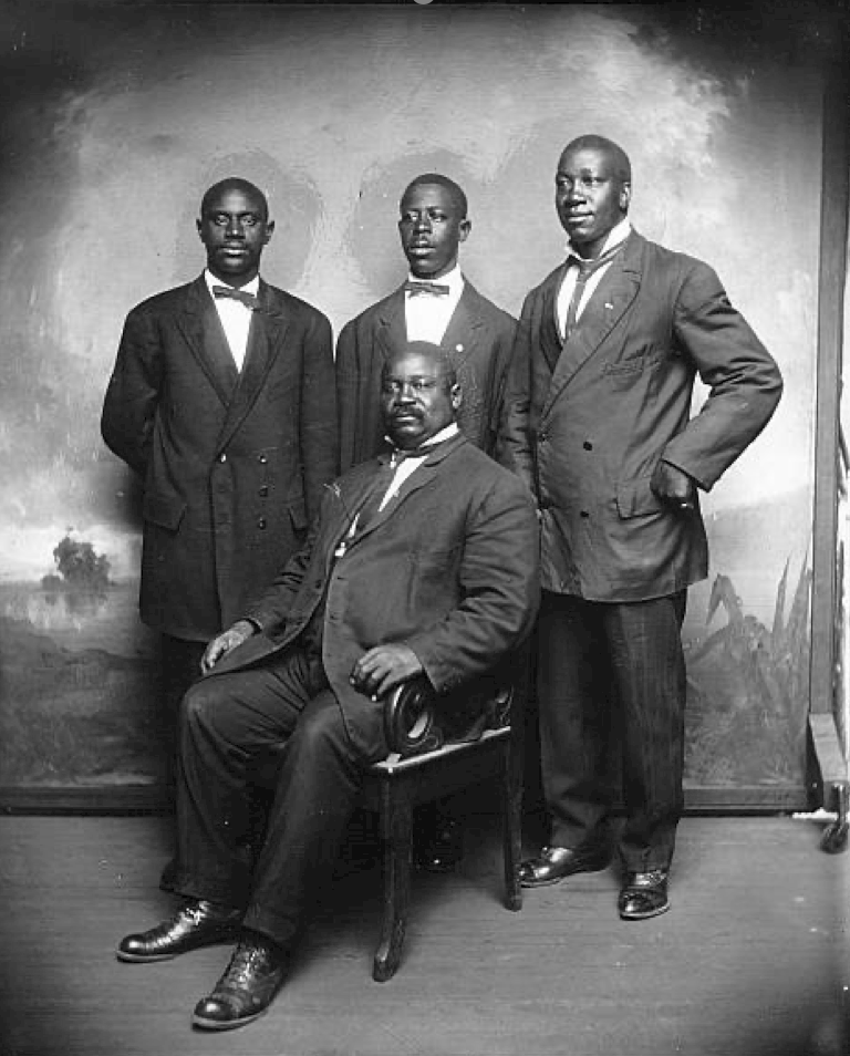 Four men in formal suits, one seated and three standing behind, posing for a group portrait in front of a painted backdrop.