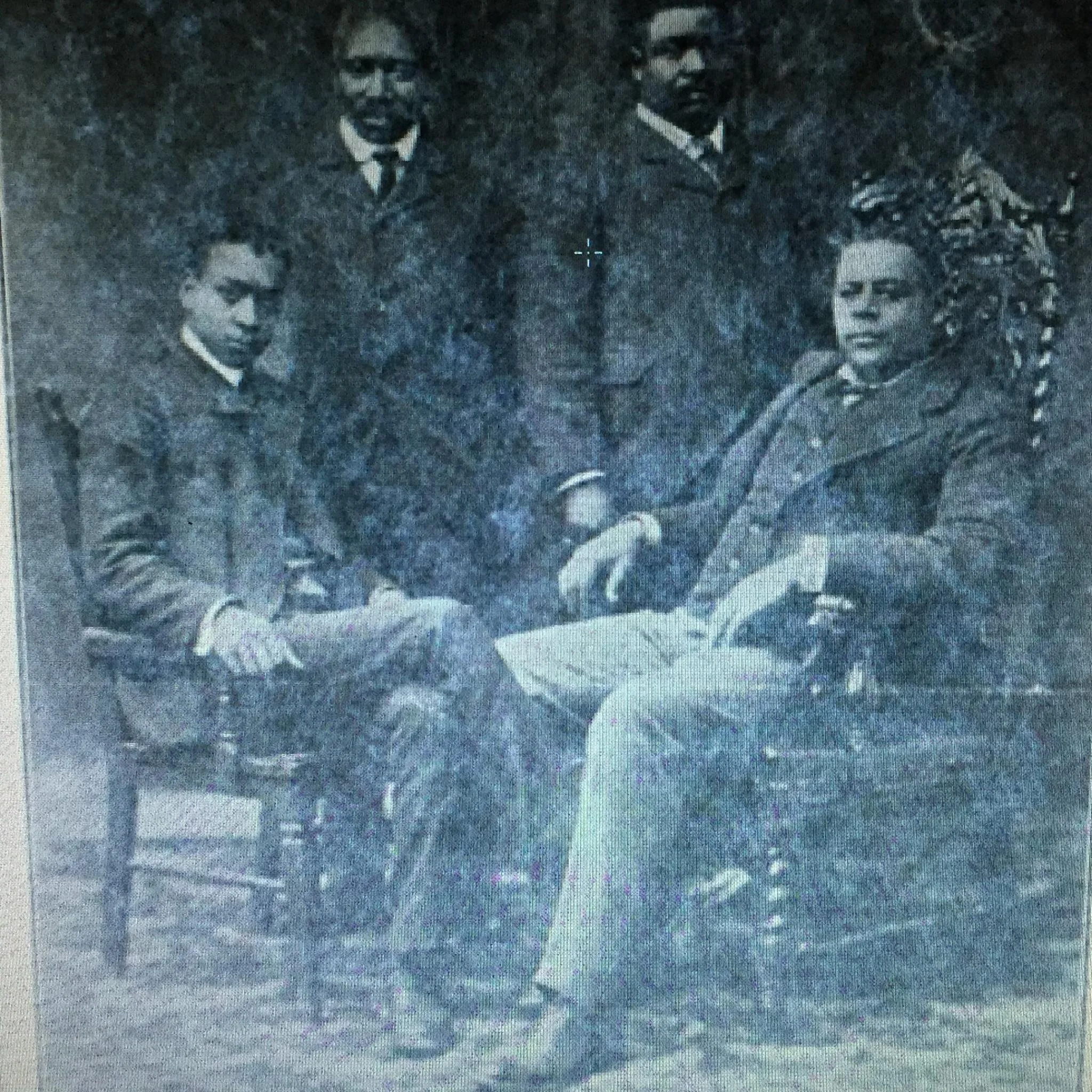 A black and white photograph of four historically dressed African American individuals, two men seated and two men standing behind them, one woman seated, with a plant in the background.