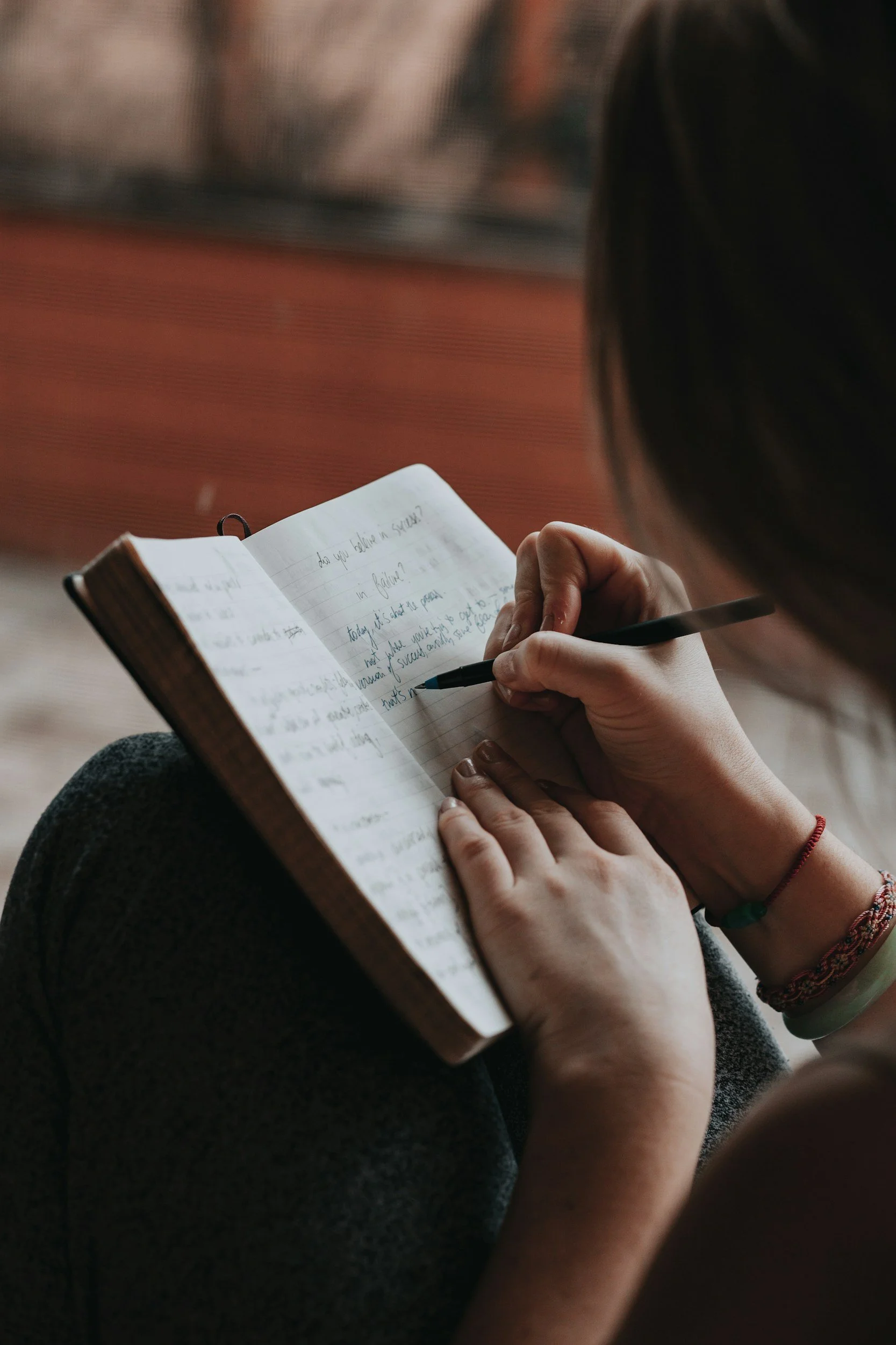 Person writing in a notebook with lined paper, sitting on their lap, with focus on hands, notebook, and pen, in a relaxed setting.