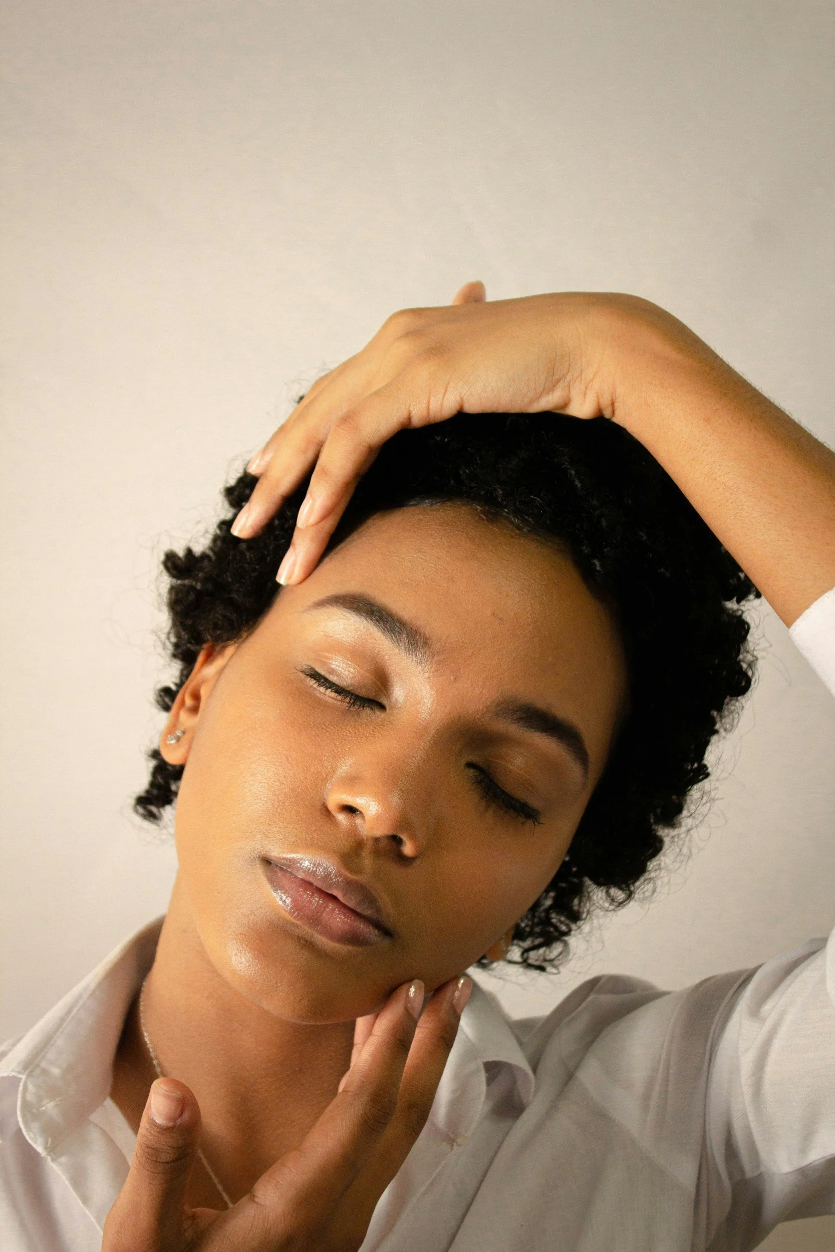 A woman with curly hair, closed eyes, and natural makeup, resting her head and hand on her forehead while gently touching her face and chest.