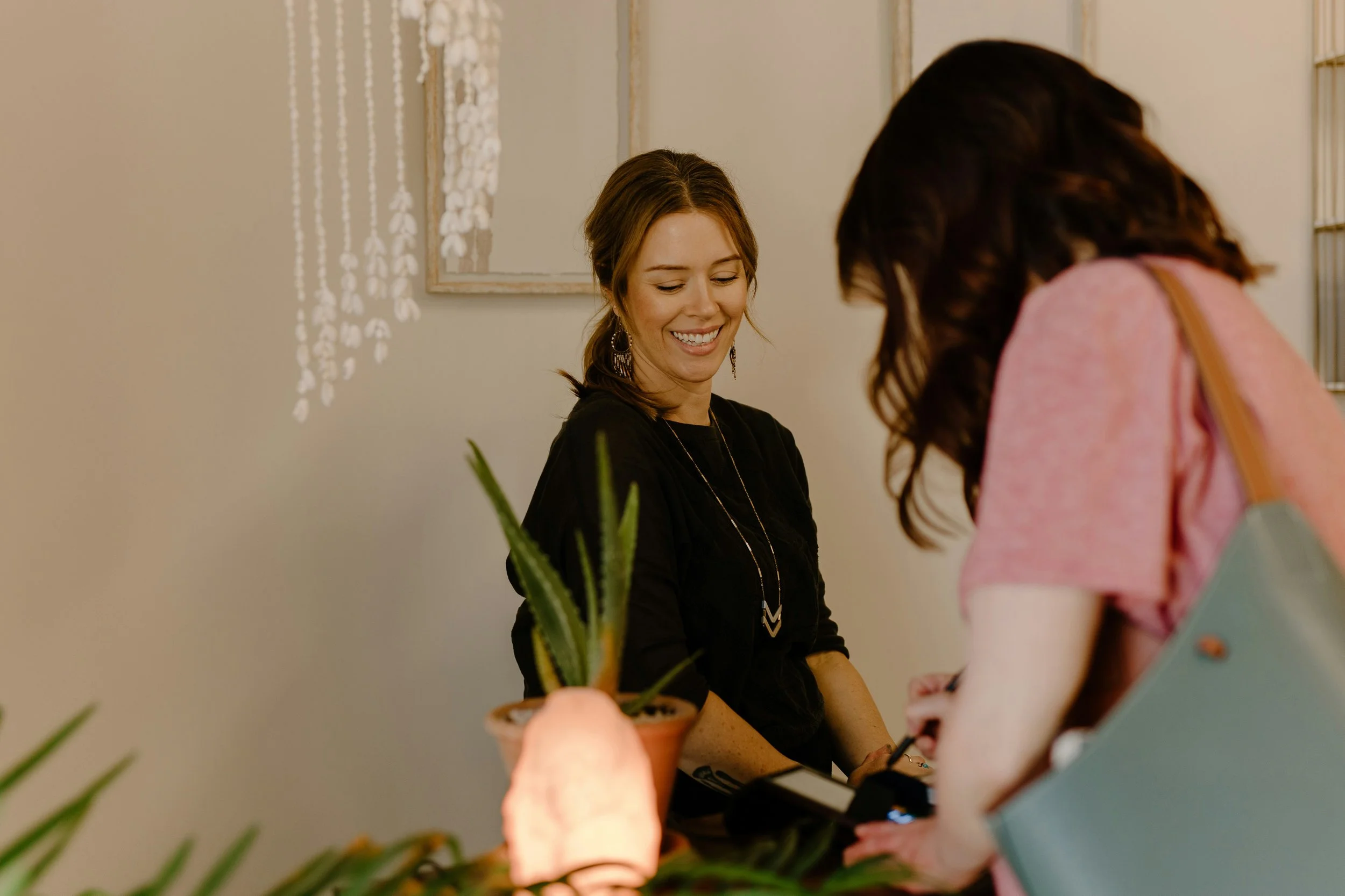 A woman sitting at a table smiling and looking down, with two other women standing nearby, one with long dark hair and the other partially visible, in an indoor setting with a light-colored wall and framed art.