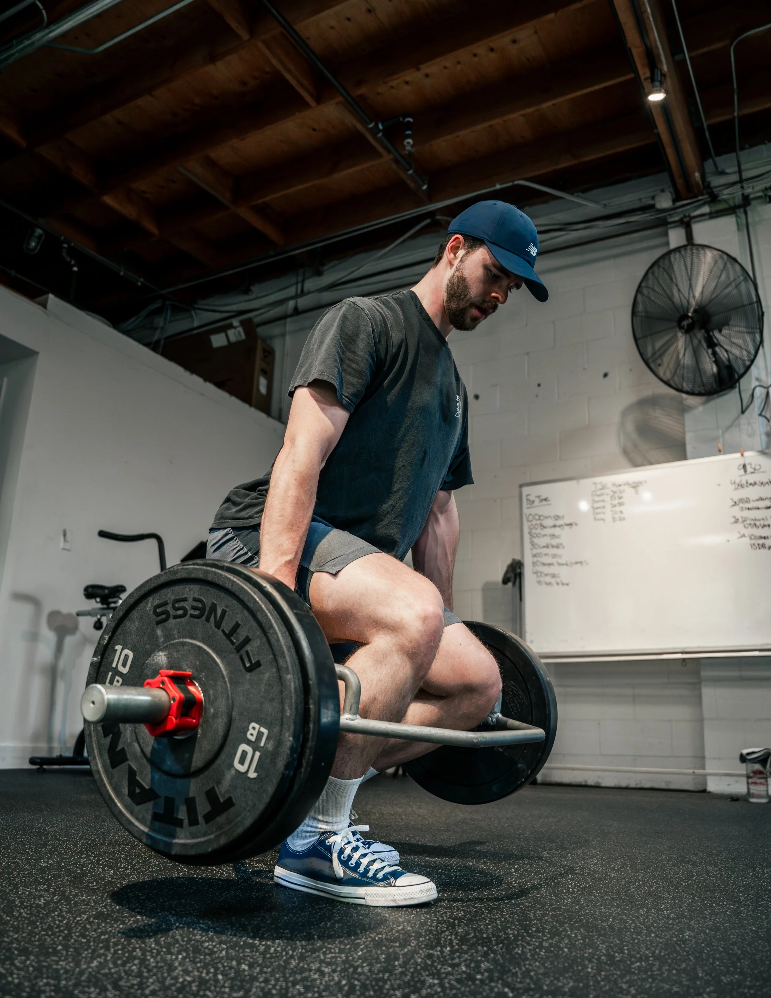 Group of four people in gym posing playfully, flexing muscles, wearing casual athletic wear. Background includes gym equipment like weights and a squat rack.