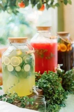 Three glass jars with fruit-infused water and drinks, surrounded by greenery and outdoor background.