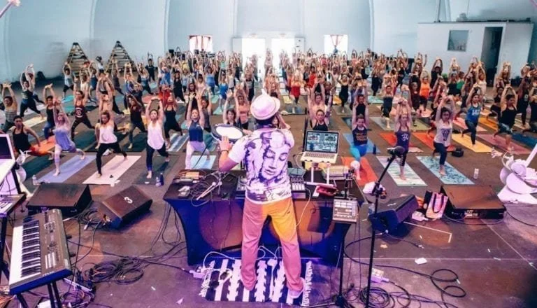 A large indoor yoga class with numerous participants on yoga mats, following an instructor on stage with musical equipment, in a spacious hall with high ceilings.