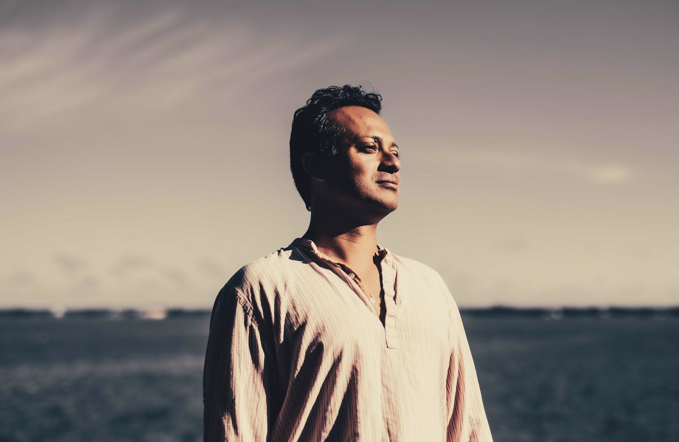 A man standing outdoors near the water, looking into the distance with a contemplative expression at sunset or sunrise.
