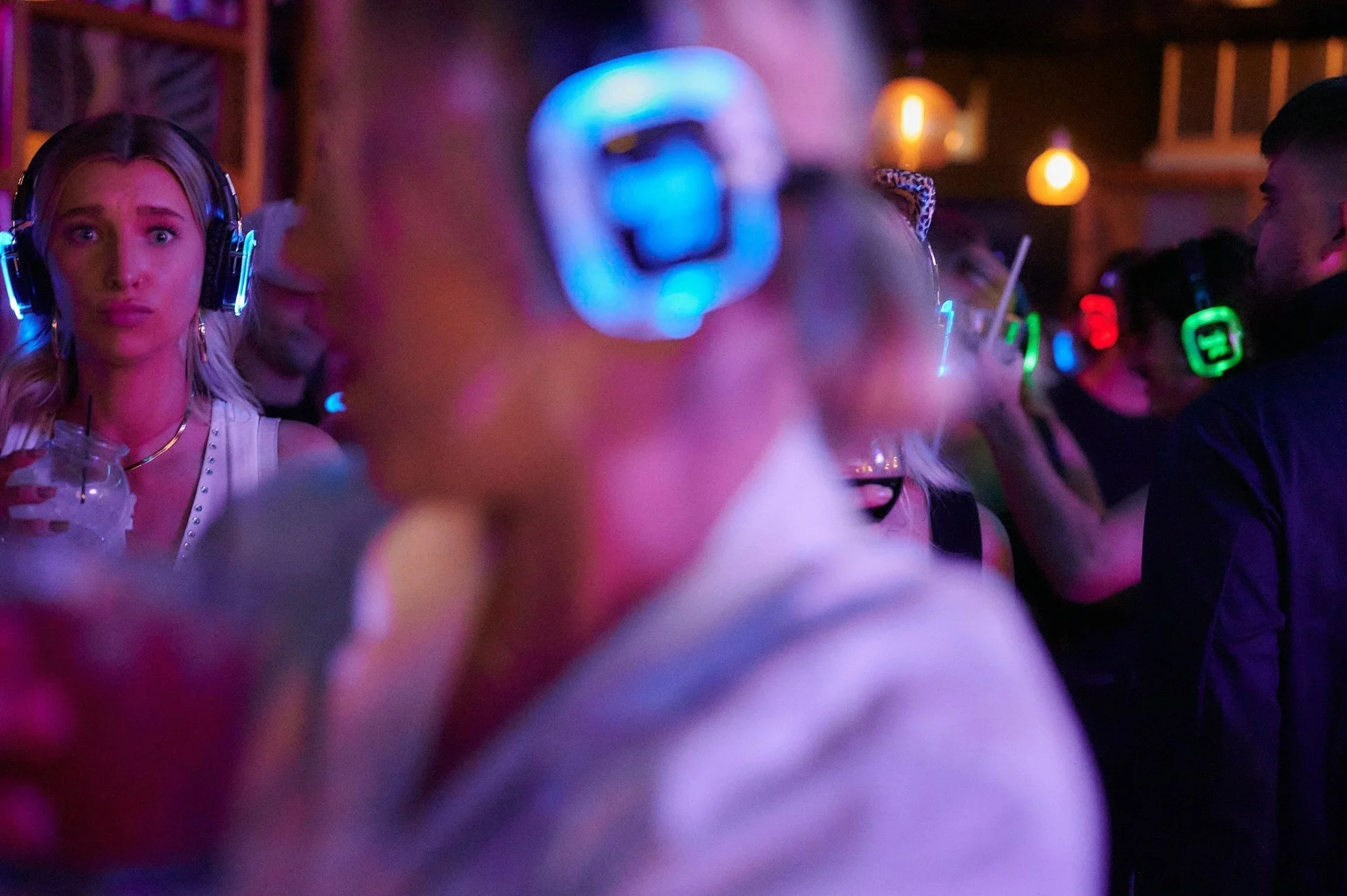 A woman with long blonde hair and headphones looks surprised or confused at a party with colorful neon lights, surrounded by other people wearing headphones in a dimly lit setting.