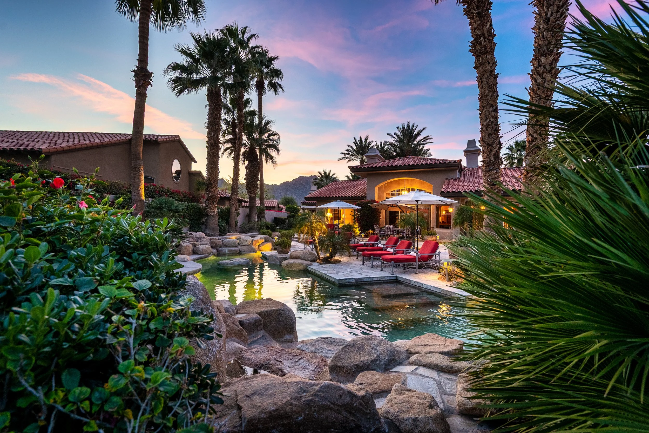 Luxury backyard with a swimming pool, palm trees, lounge chairs with red cushions, and a house with outdoor lighting at dusk.