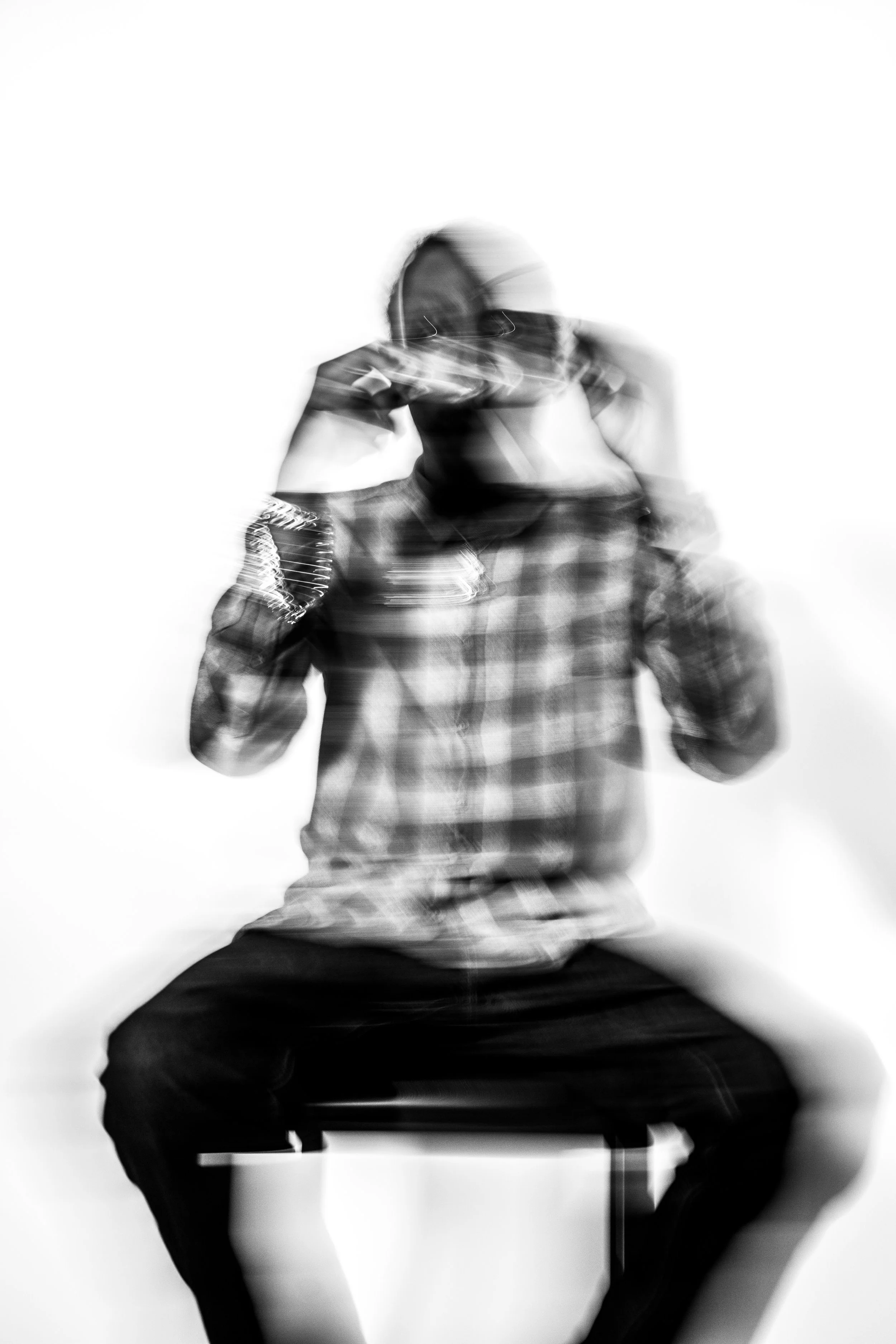 Blurred black and white photo of a person sitting on a stool, wearing sunglasses and a checkered shirt, adjusting their glasses with both hands.