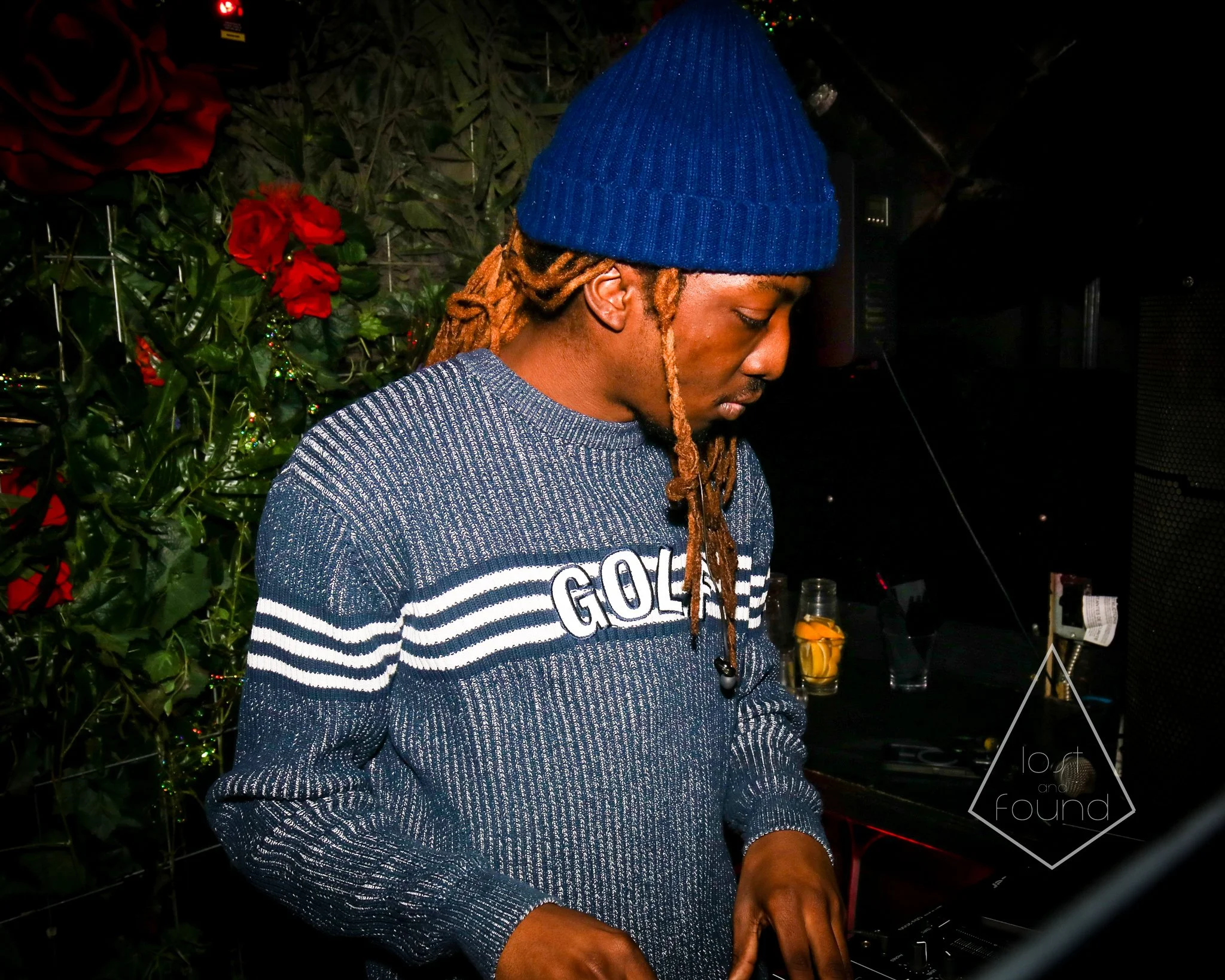A man with long, dreadlocked hair wearing a blue knit beanie, a blue and gray sweater with white stripes and the word 'GOLF' on it, appears focused while DJing in a dimly lit room with red roses and green foliage in the background.