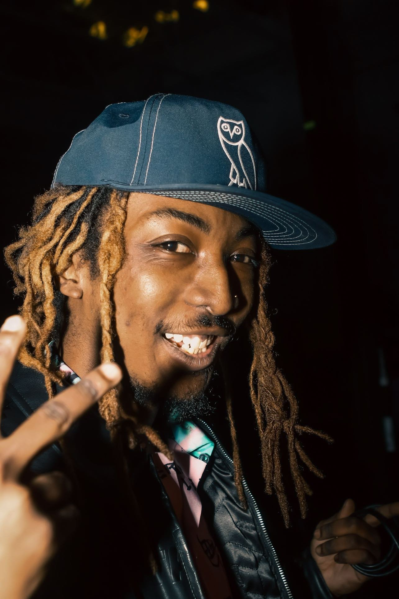 A man with dreadlocks wearing a blue cap with an owl logo, smiling, and making a peace sign with his fingers.