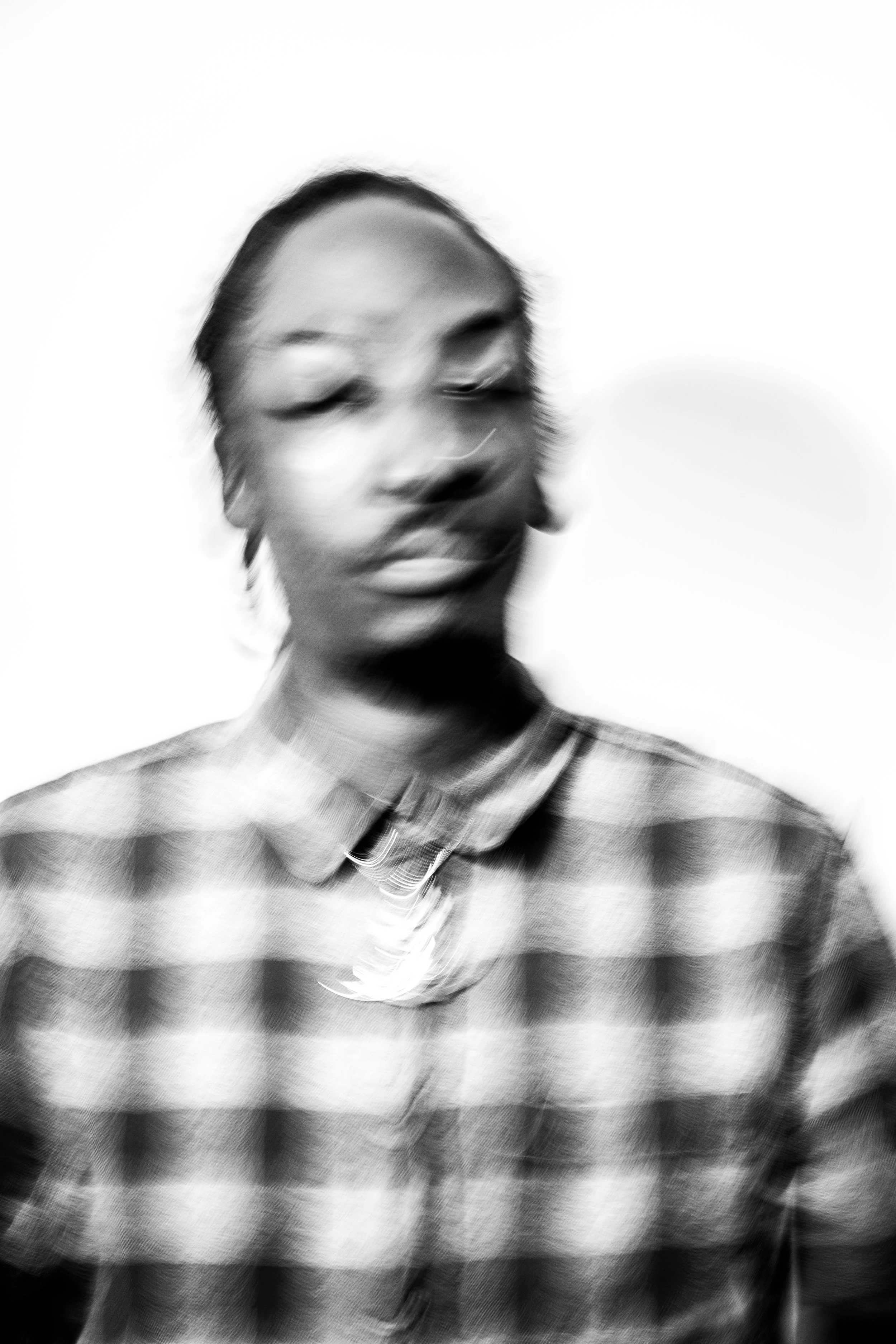 A black and white portrait of a person with blurred facial features, wearing a plaid shirt and layered necklaces in a minimalist style.