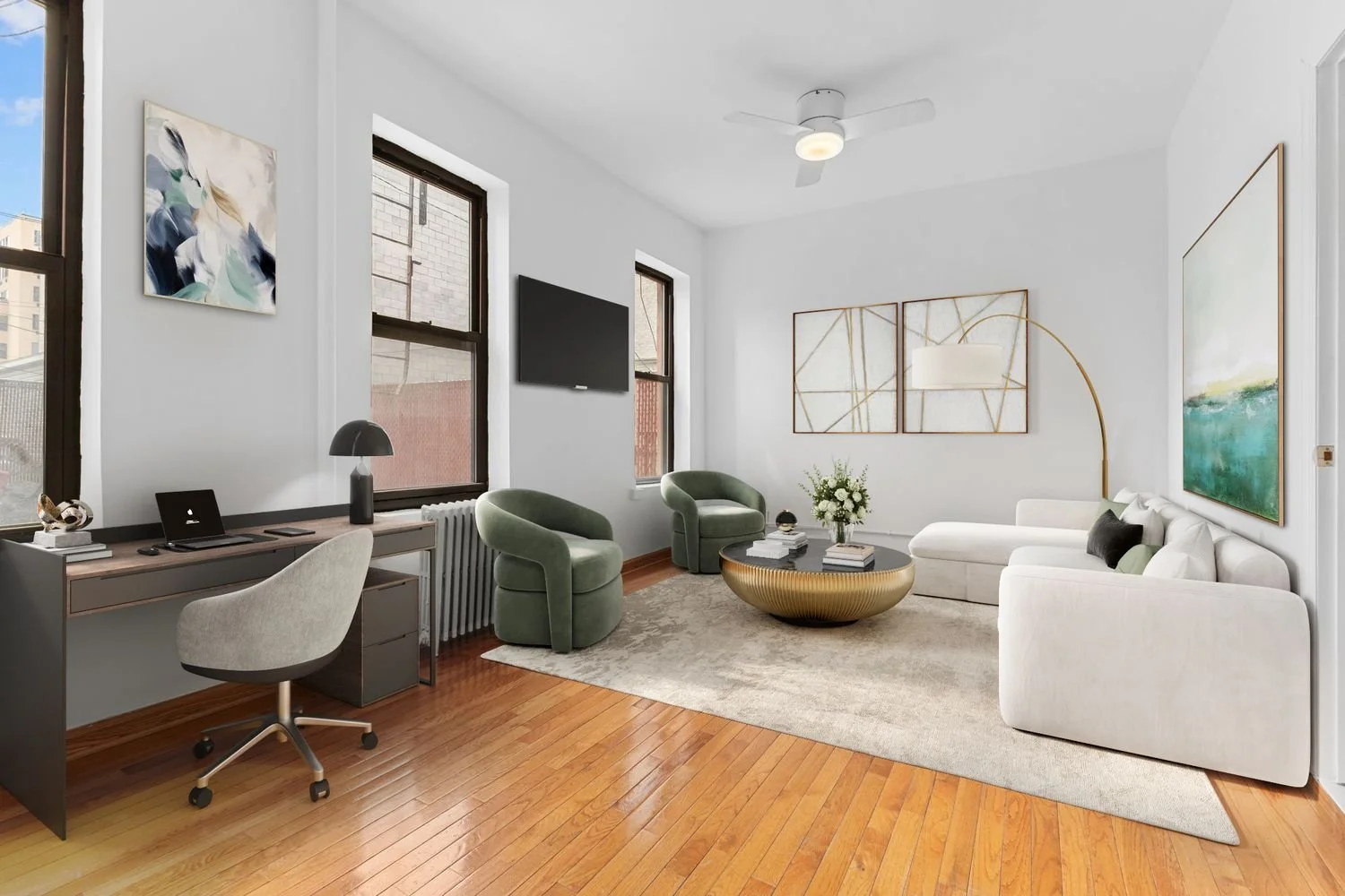 A modern, sunlit living room featuring white walls and medium-tone hardwood floors. On the left, a sleek dark wood desk with a grey swivel chair sits beneath a window.