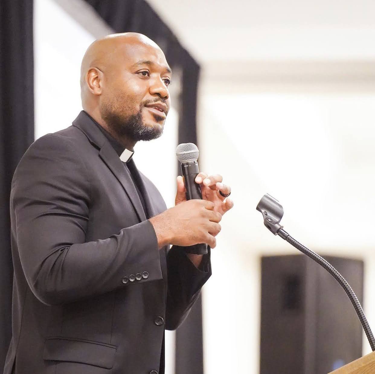 Meshach Kanyion wearing a black suit and clerical collar speaking into a microphone at a podium.