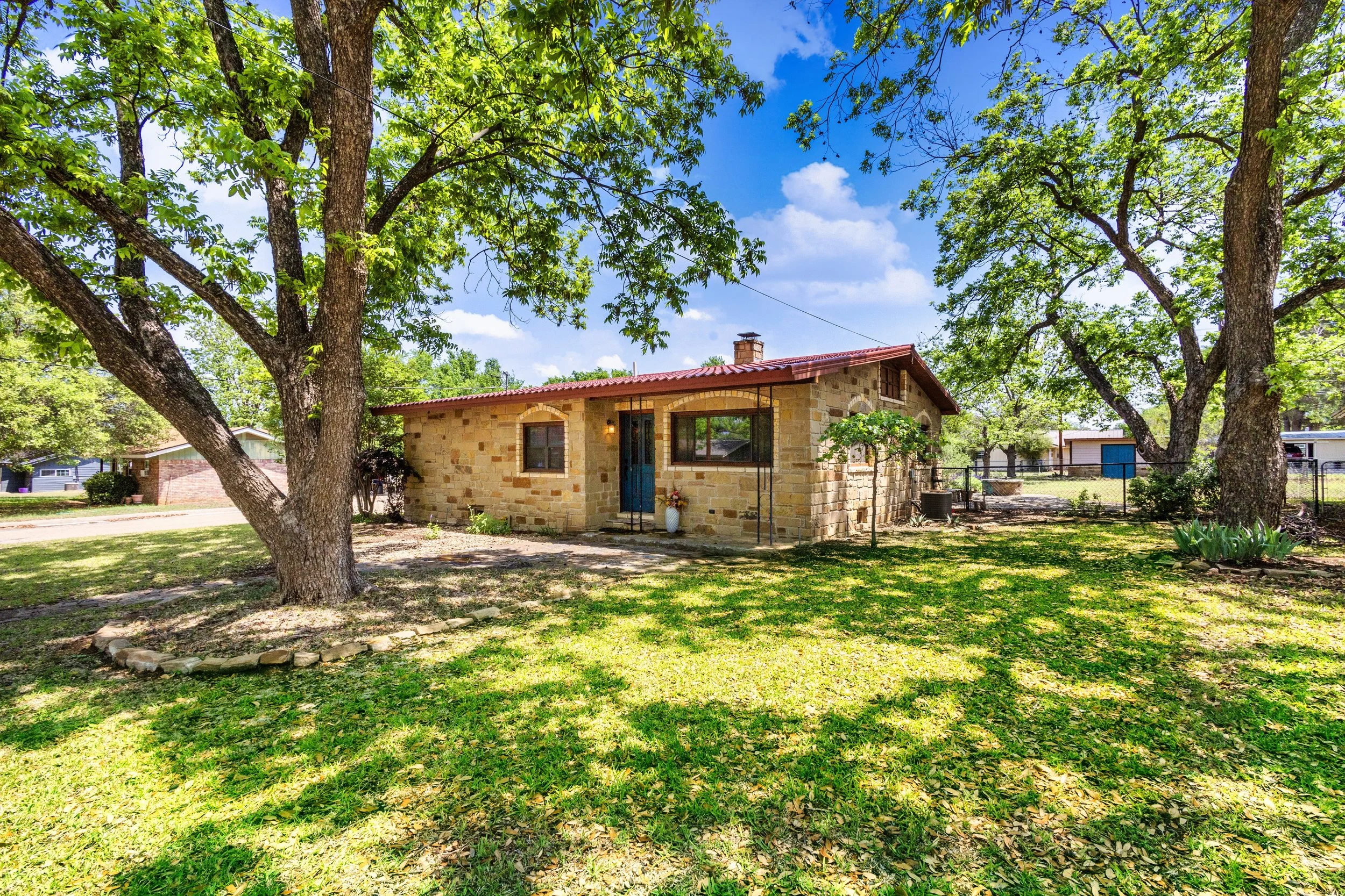 A small stone house with a red roof, blue front door, and large windows, surrounded by green trees and a grassy yard on a sunny day.
