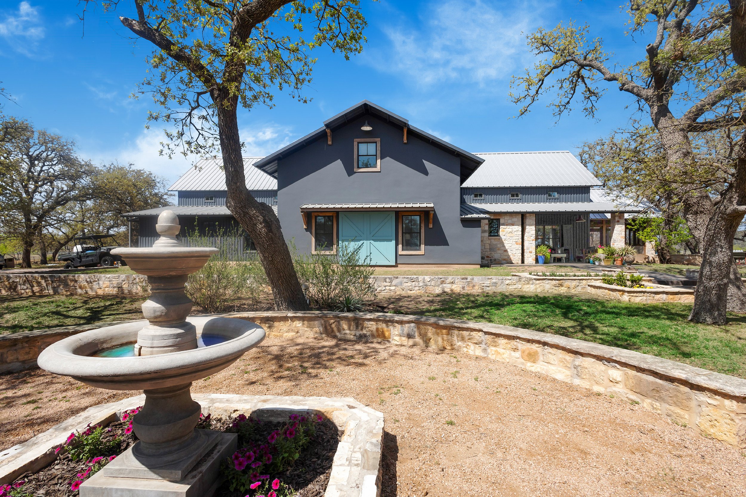 A large gray house with a blue door, stone accents, and a metal roof, surrounded by trees and a landscaped yard, with a fountain in the foreground under a blue sky.