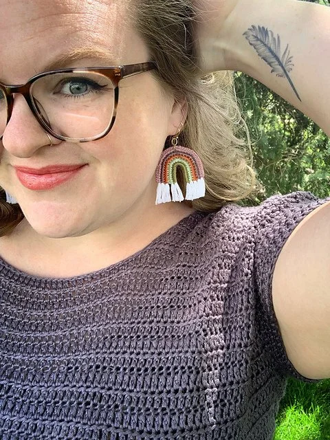 A woman with glasses holds her long blond hair back to show the crocheted rainbow earring she is wearing.