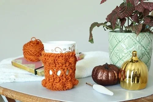 A coffee cup with an orange cup cozy sits on a table with fall decorations, a ball of yarn, a crochet hook and a plant in a green textured pot.