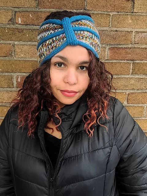 A woman with dark curly hair wears a headband-style earwarmer crocheted with blue and variegated yarn.