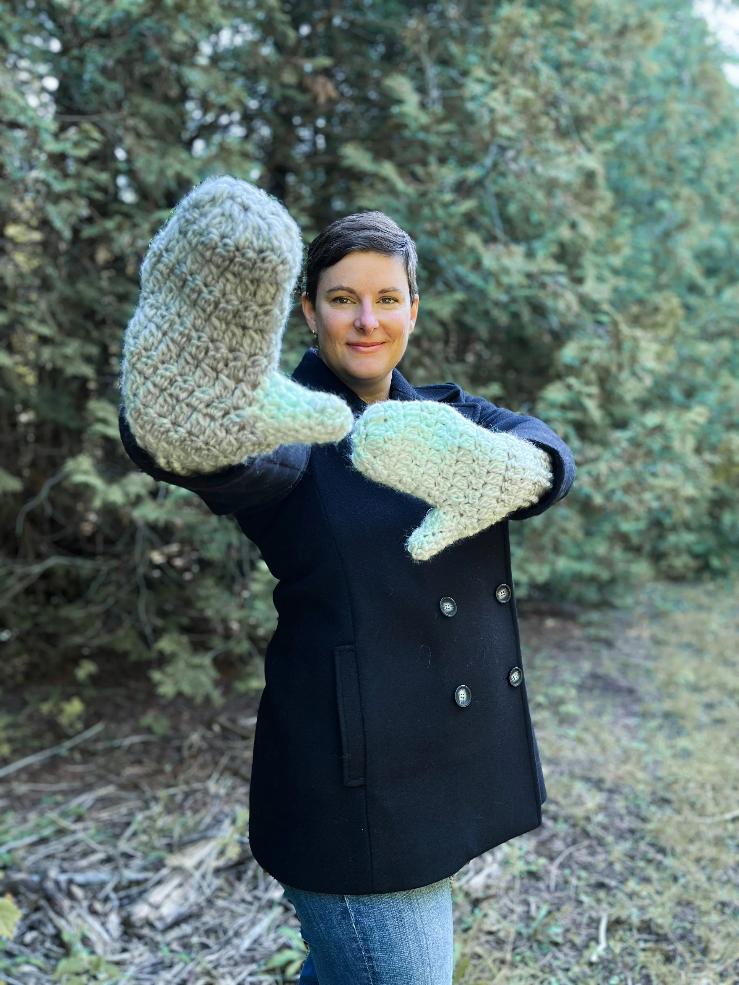A woman with short hair in a black wool peacoat holds her hands in front of her to show off the mint green and grey mittens she is wearing.